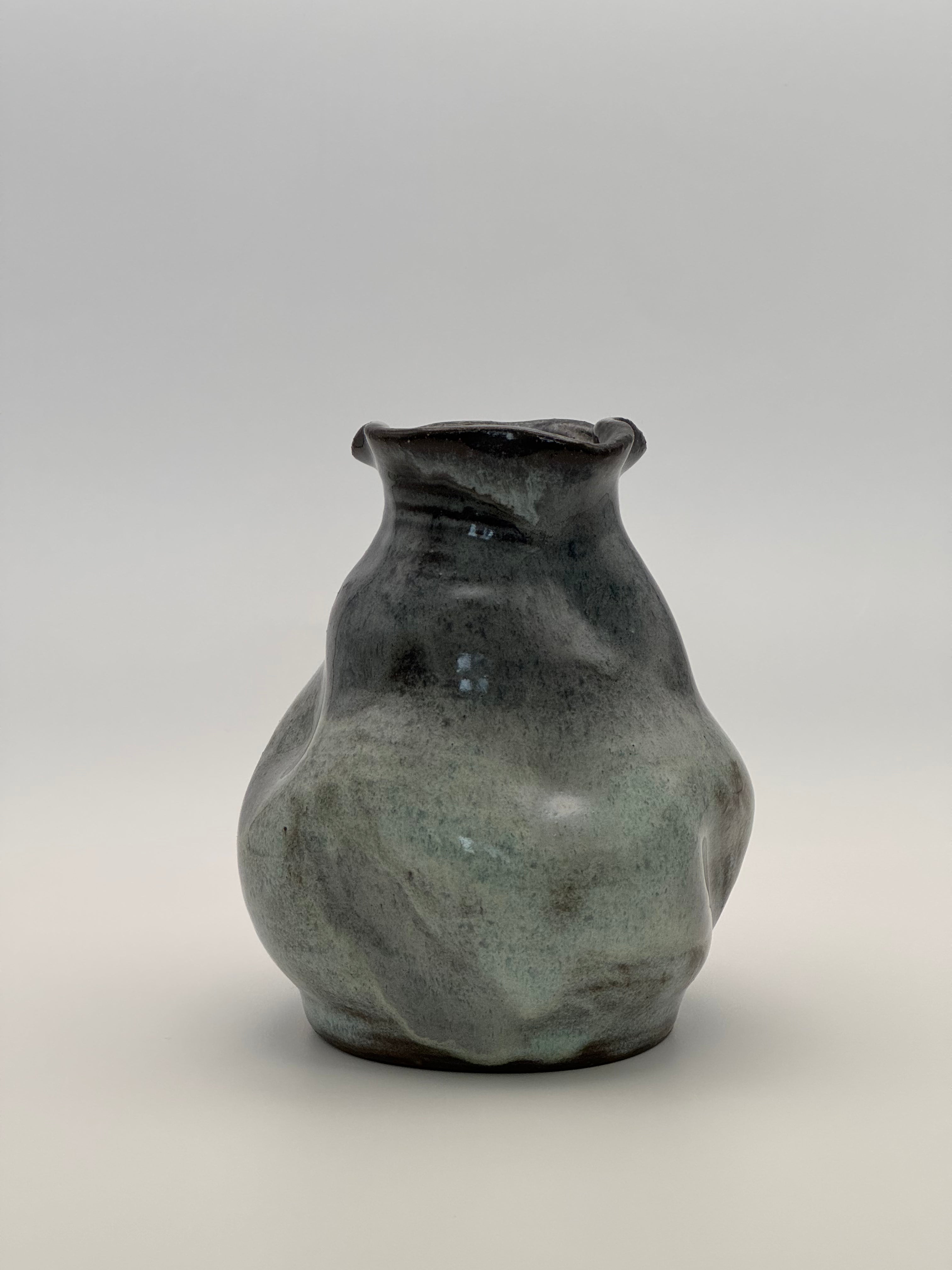 A handmade ceramic vase with a rounded, organic shape and a small flared rim, featuring a mottled glaze that transitions from dark blue to light blue, set against a neutral gray background.