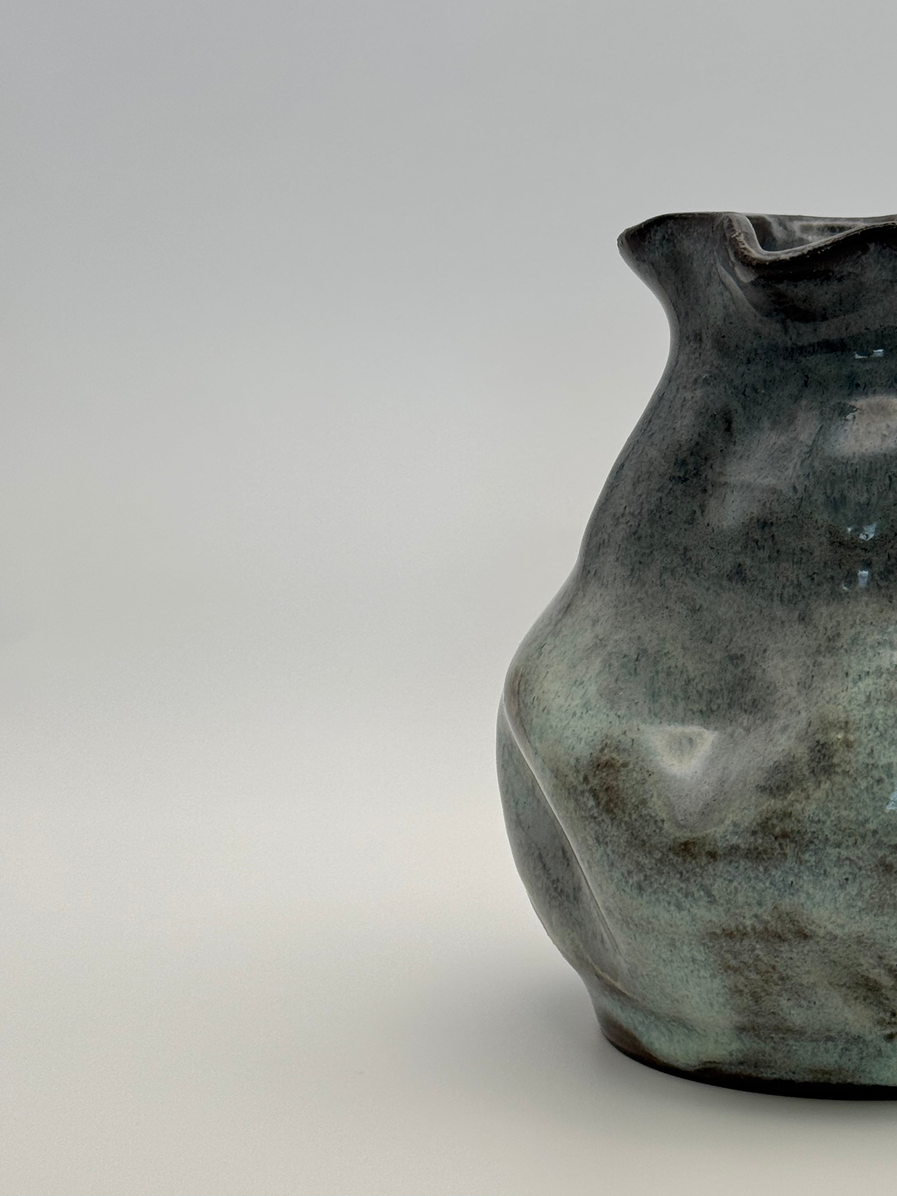 A close-up studio shot of A handmade ceramic vase with a rounded, organic shape and a small flared rim, featuring a mottled glaze that transitions from dark blue to light blue, set against a neutral gray background.