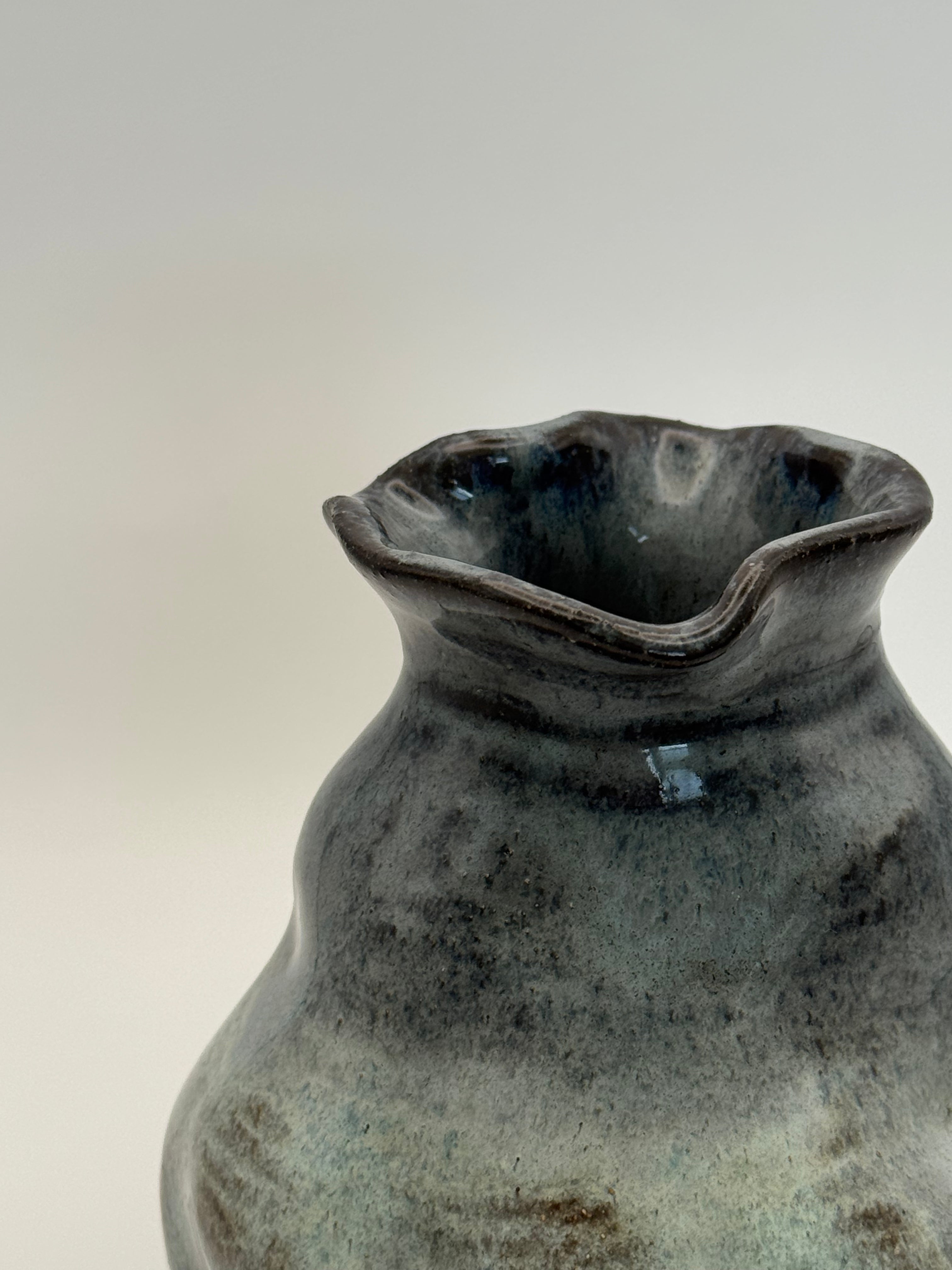 close up of the wavy rim of a  handmade ceramic vase with a rounded, organic shape and a small flared rim, featuring a mottled glaze that transitions from dark blue to light blue, set against a neutral gray background.