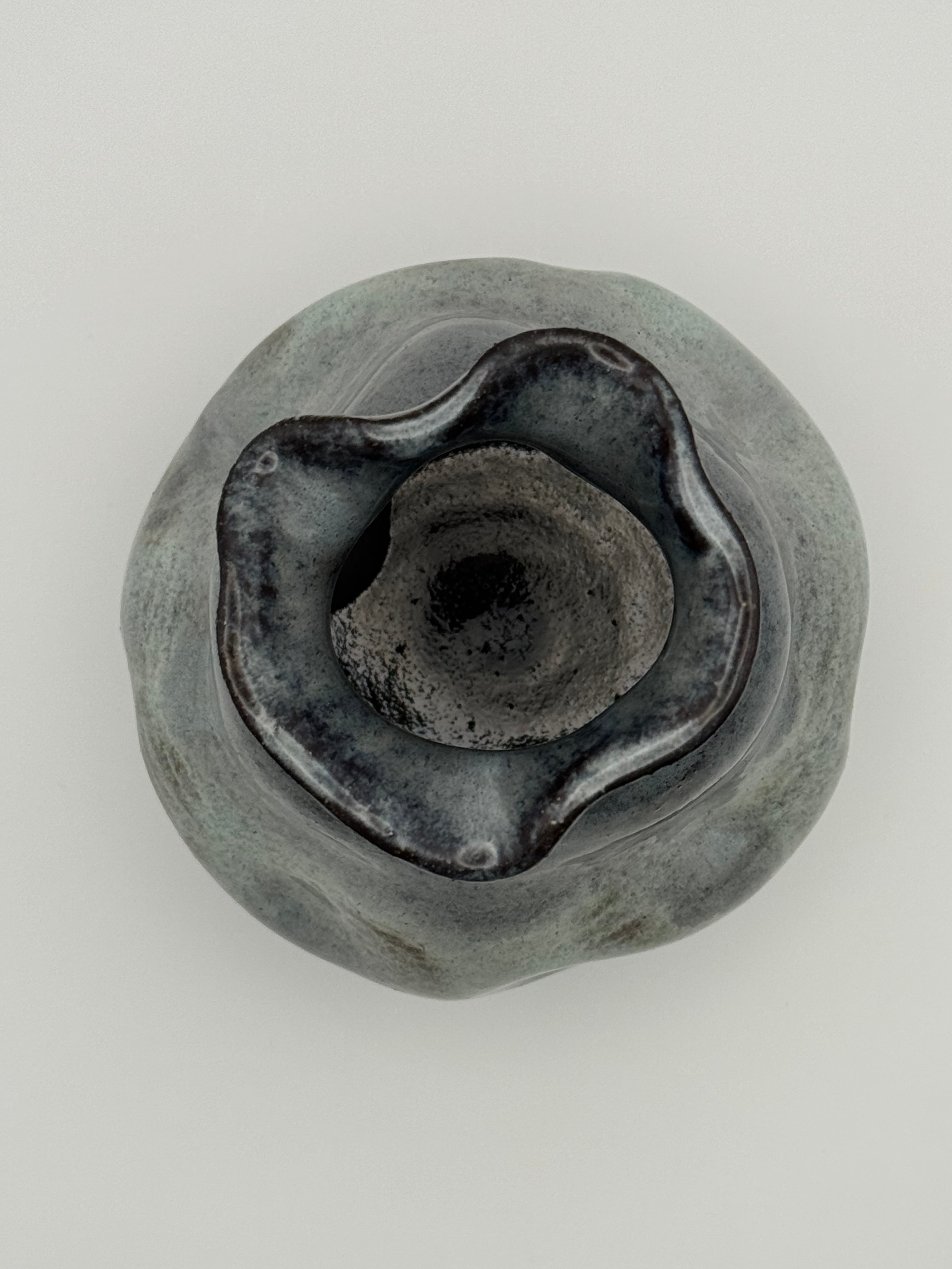 A top-down view of A handmade ceramic vase with a rounded, organic shape and a small flared rim, featuring a mottled glaze that transitions from dark blue to light blue, set against a neutral gray background.