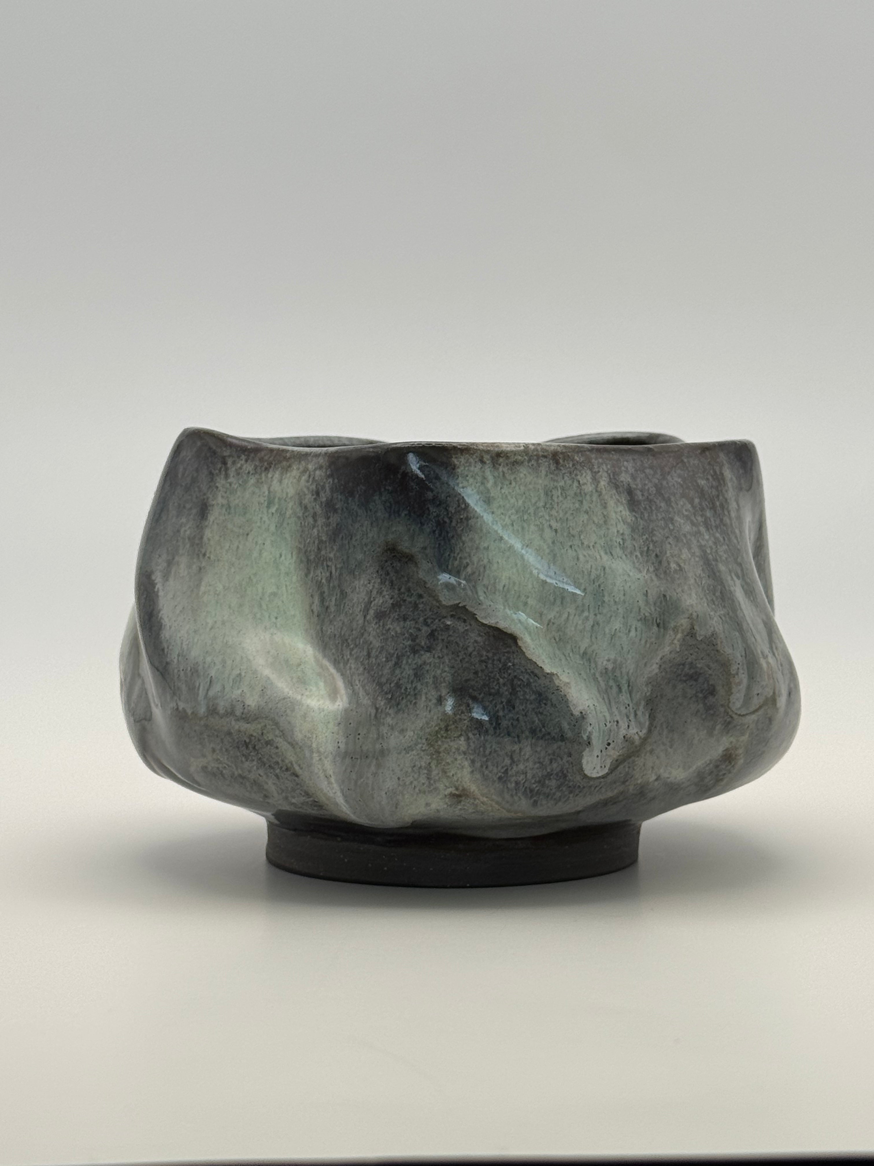 A handmade ceramic bowl with a dark, organic, wavy shape and a mottled glaze of charcoal blue-gray, black, and muted blue, sitting on a small black base against a neutral gray background.