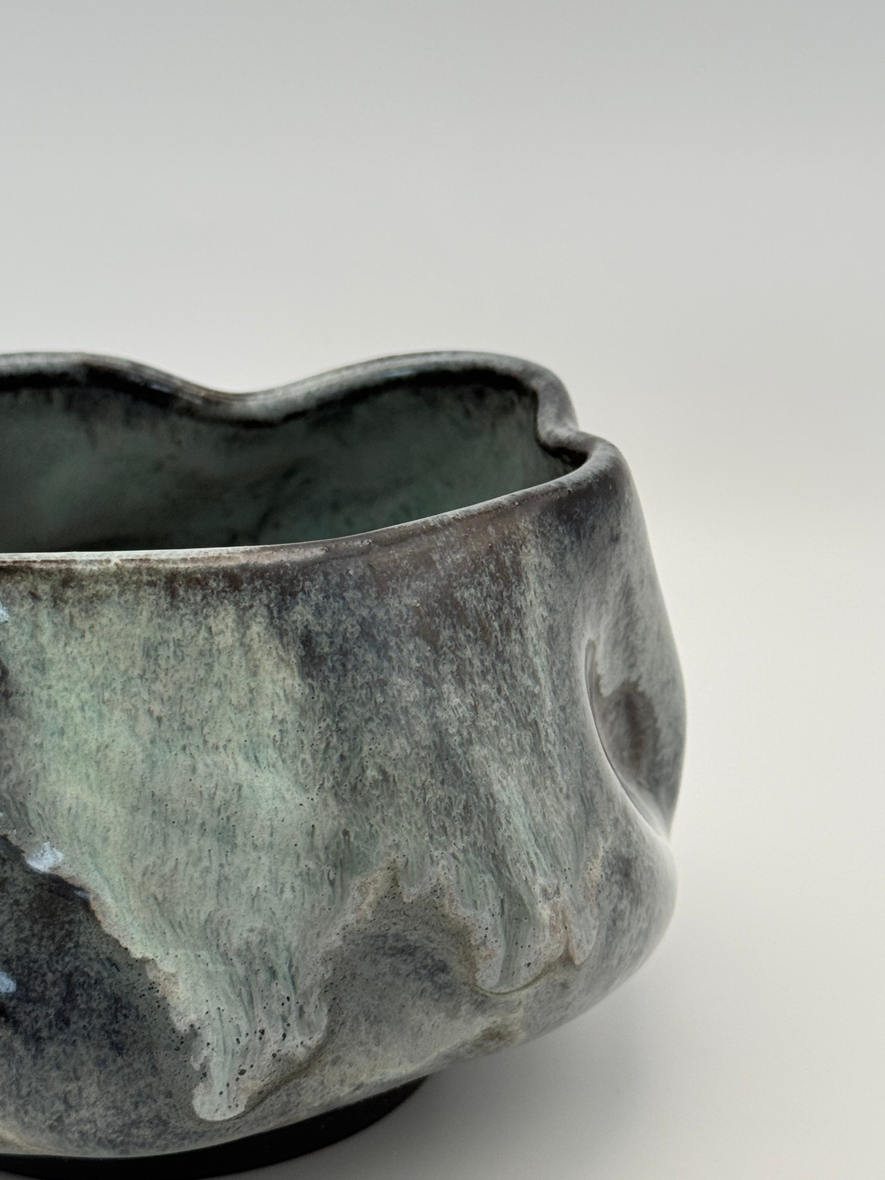 Close-up of a handmade ceramic bowl with a wavy, organic rim and a textured glaze showing dark blue, black, and muted blue tones, set against a neutral light gray background.