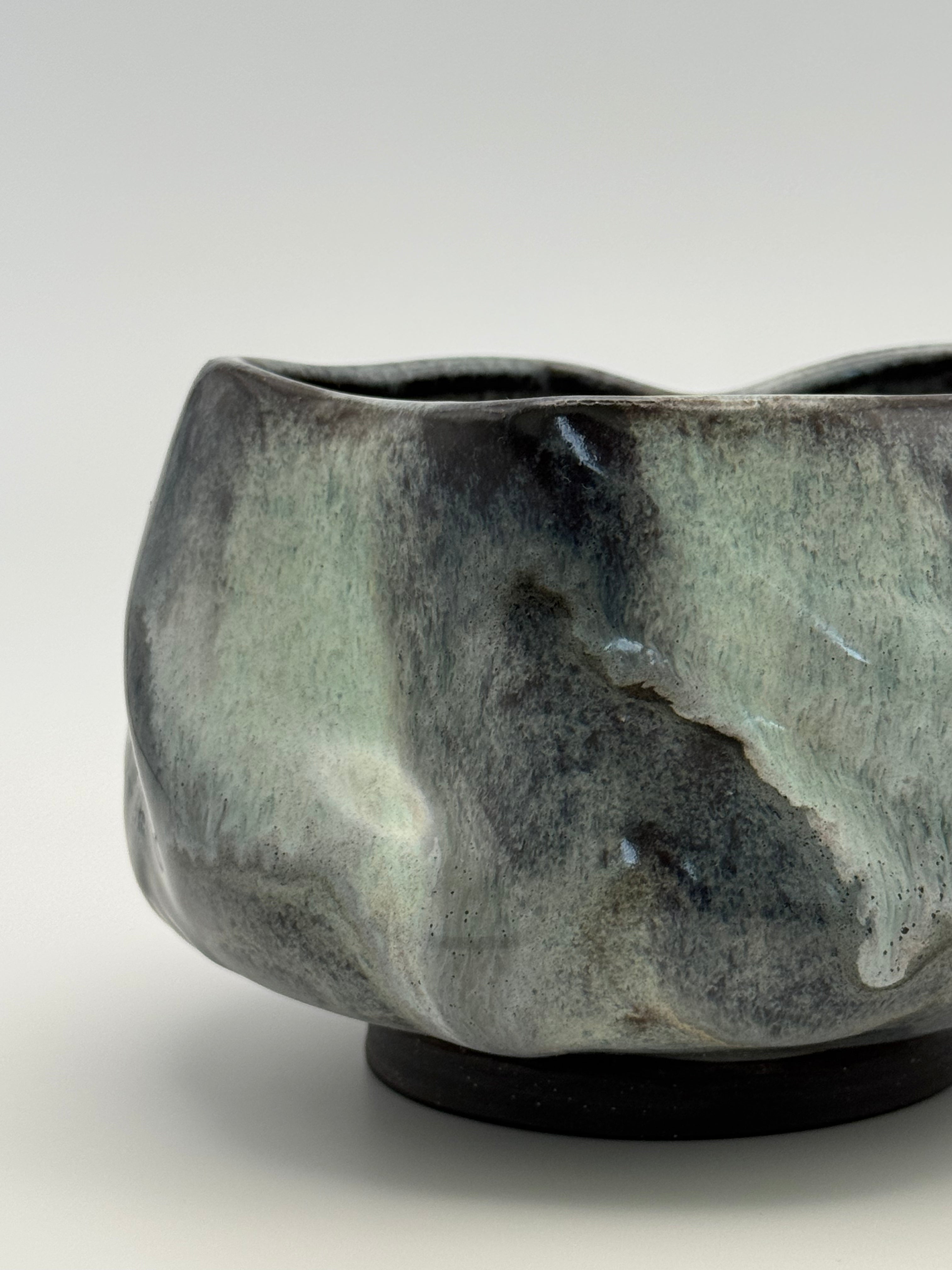 A close-up of a handmade ceramic bowl with a textured, organic shape and a mottled glaze of dark blue, black, and muted blue, sitting on a plain light gray background.