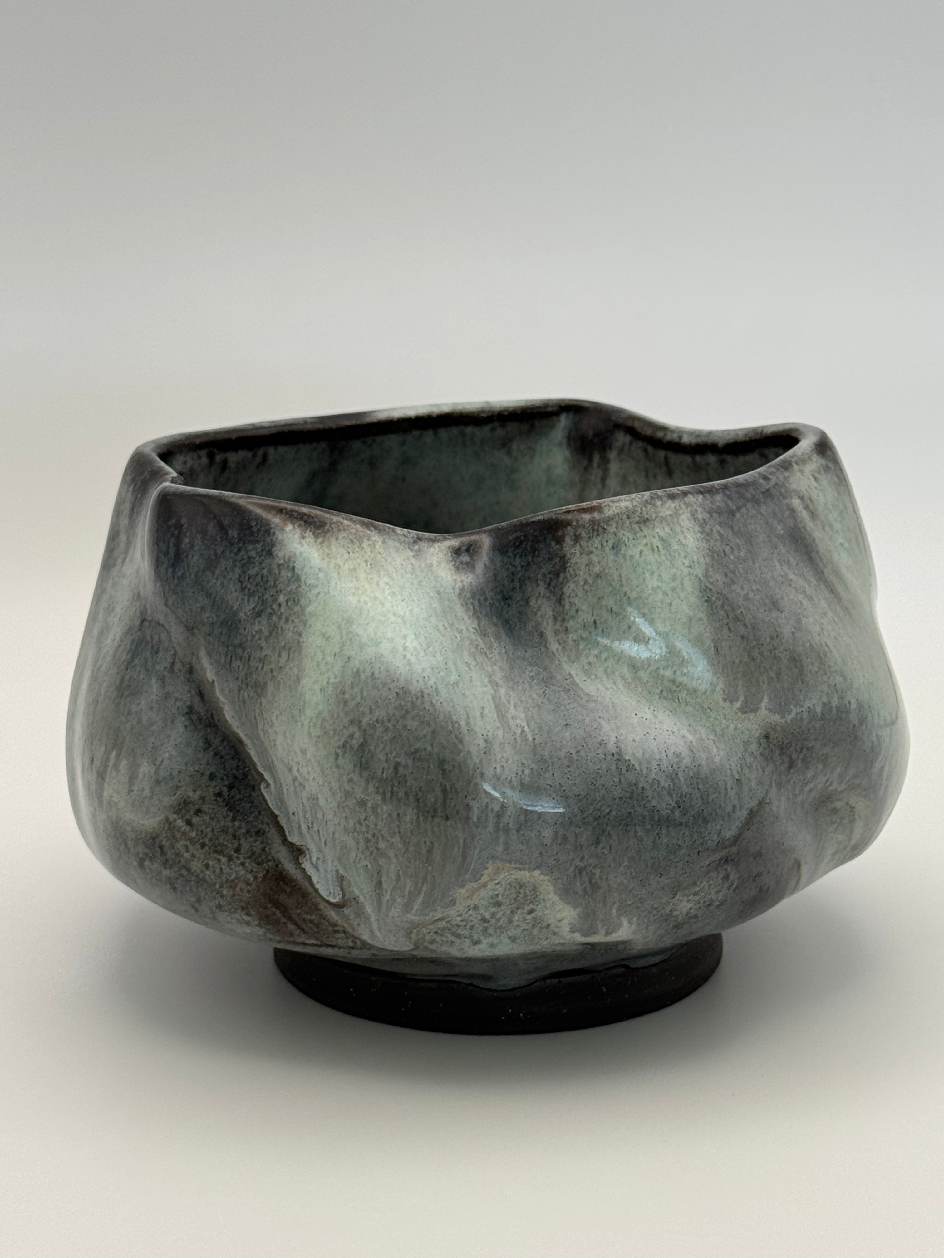 A handmade ceramic bowl with an organic, wavy silhouette and a textured glaze featuring mottled shades of dark blue grey, black, and muted blue, set against a neutral gray background.