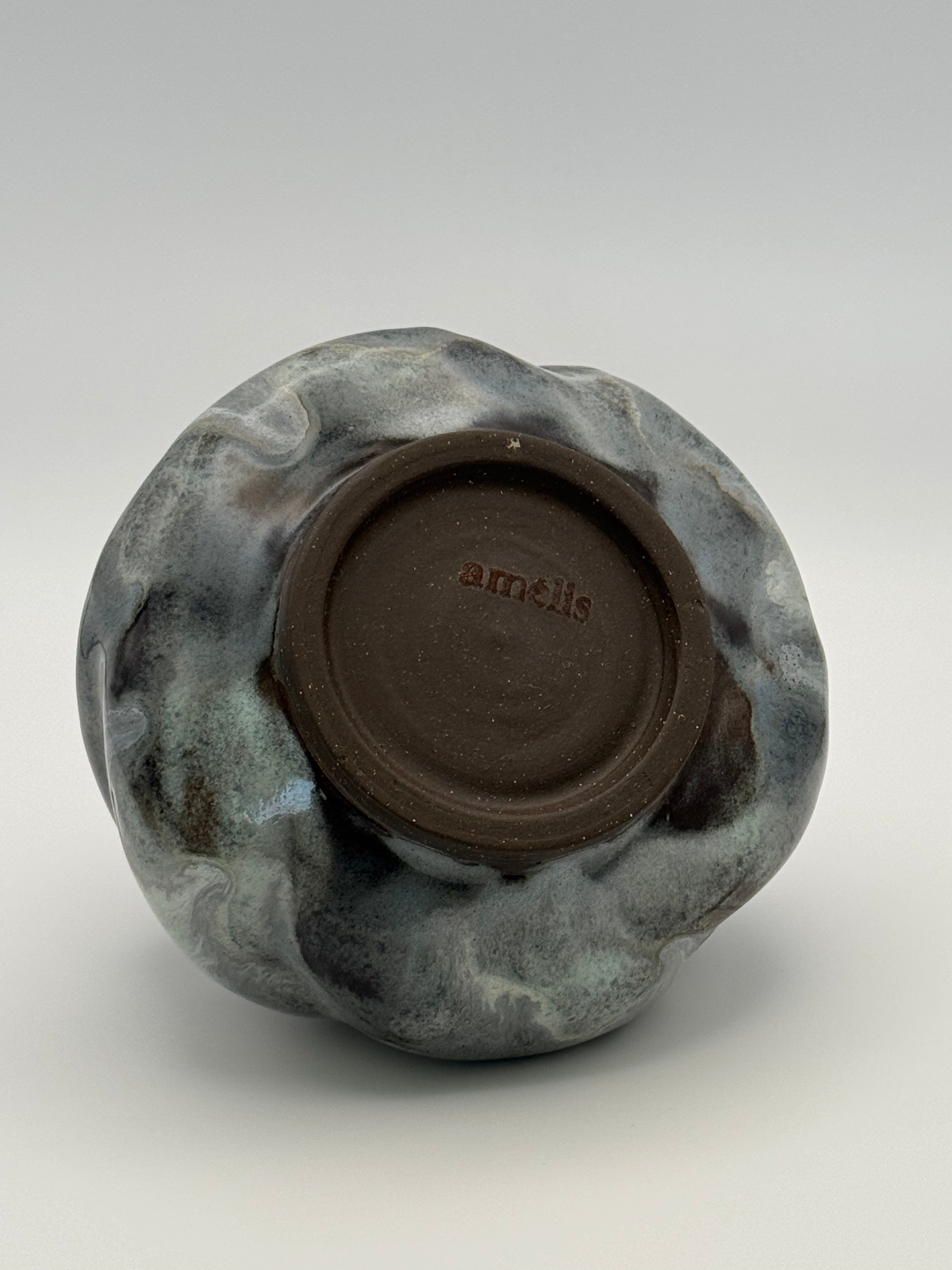 The underside of a ceramic bowl with a swirled blue-gray and white glaze, showing a dark brown clay foot ring stamped with the brand name 'amélis'.