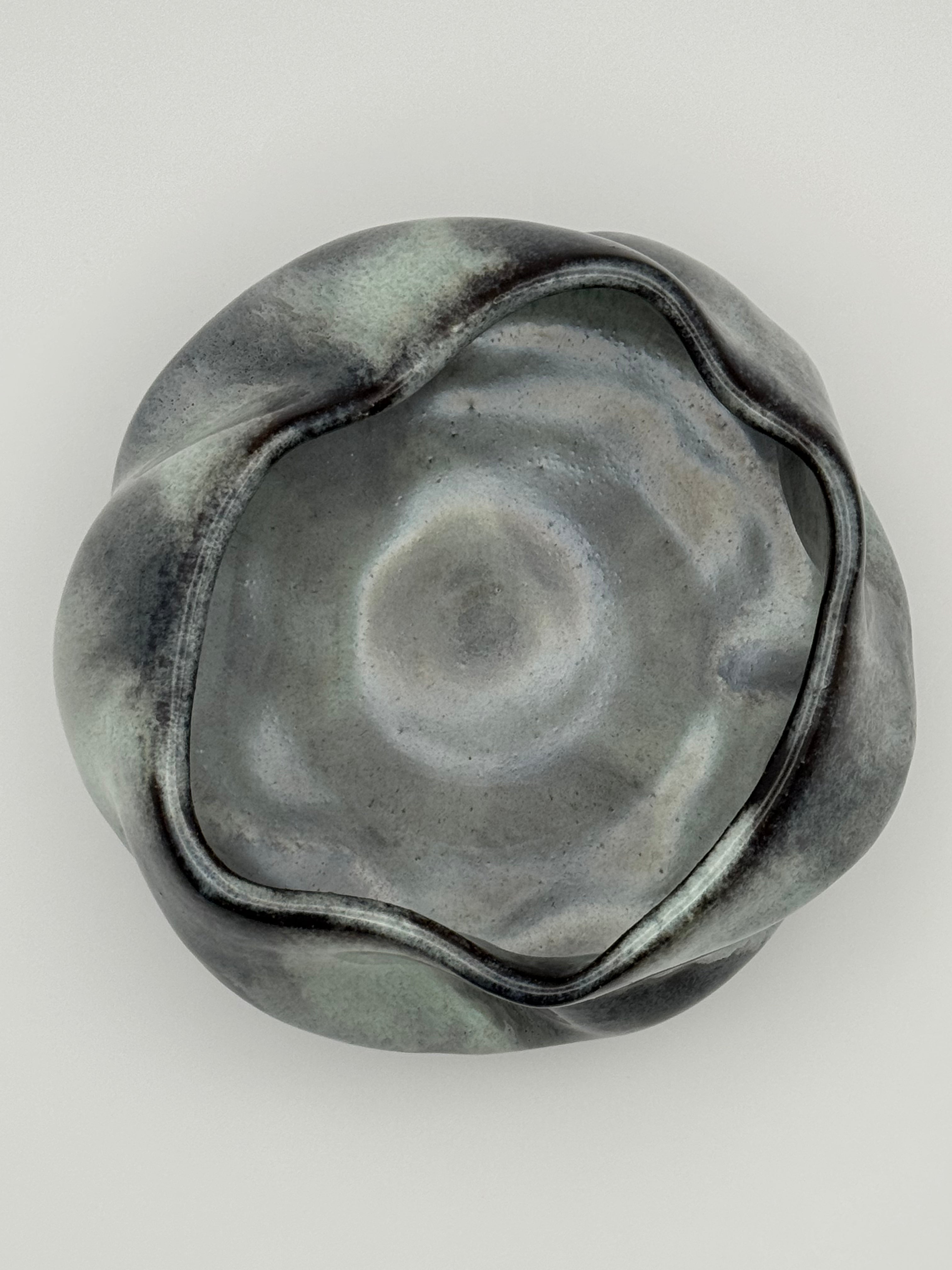 A sculptural ceramic bowl with a wavy, organic rim and a smooth, glossy interior, shown from a top-down perspective on a light gray background.