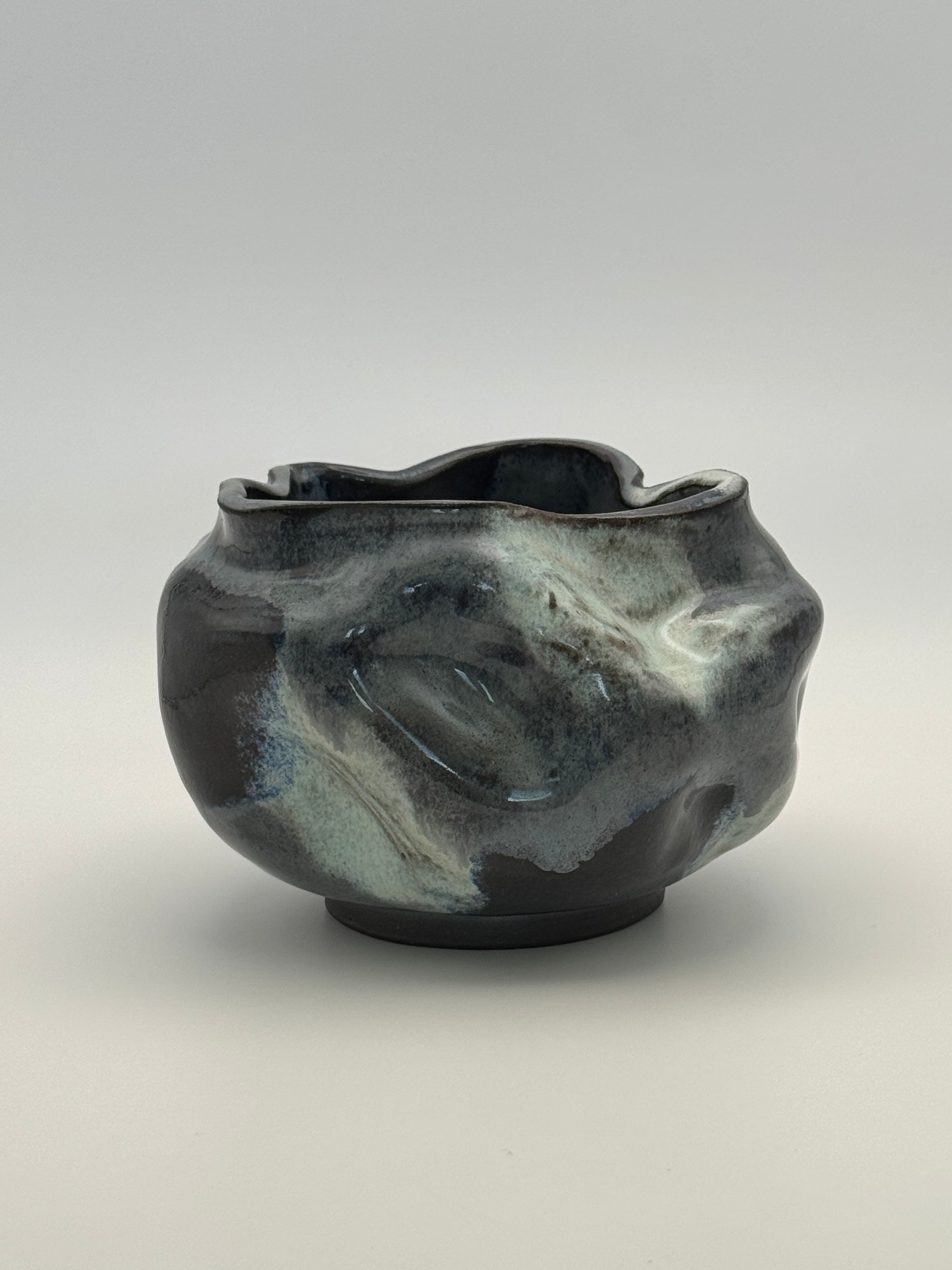 A handmade ceramic bowl with an organic, wavy shape and a glossy, marbled glaze in darkblue, blue-gray, and light blue, set against a neutral gray background.