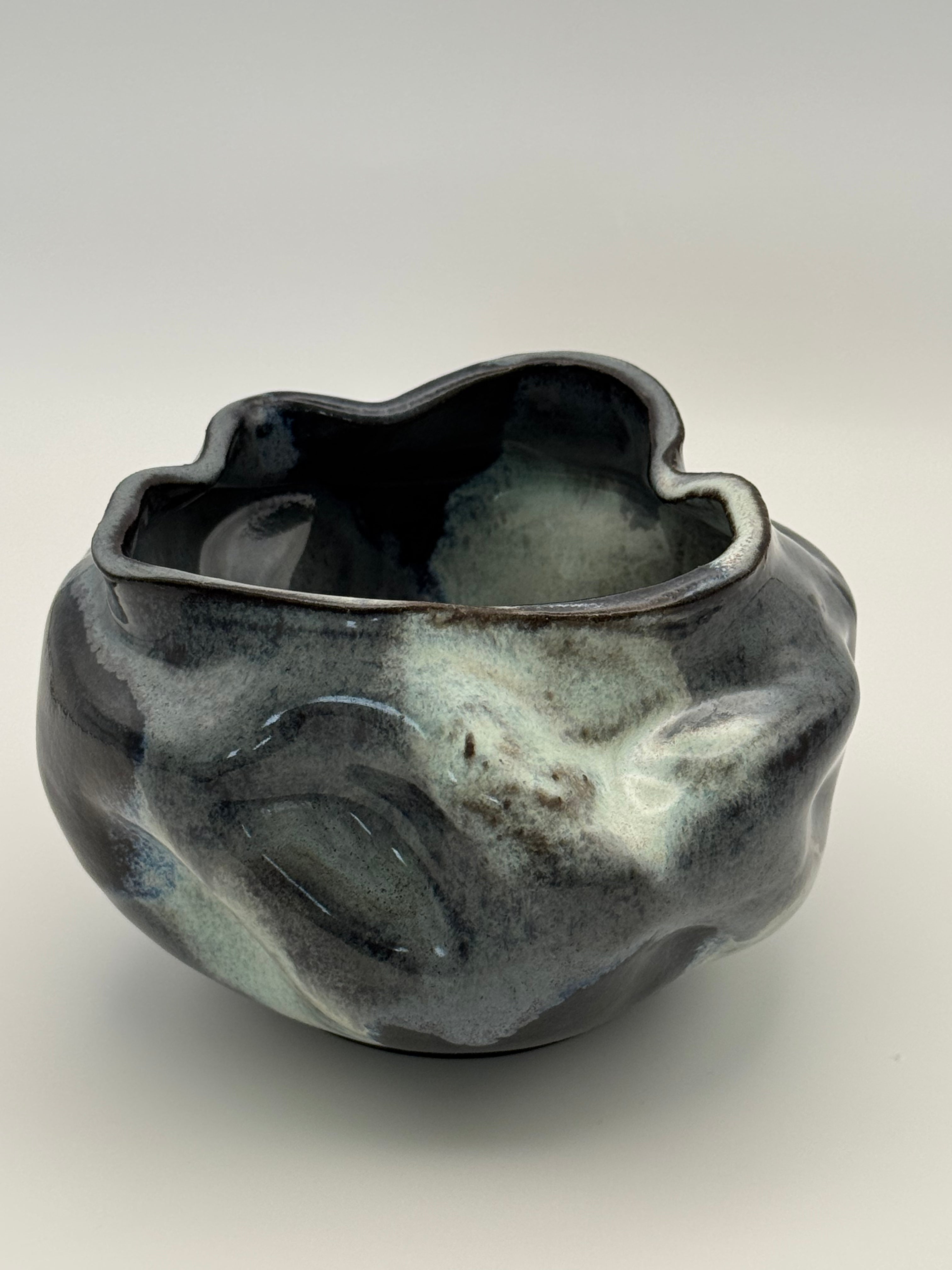 A handmade ceramic bowl with an organic, wavy shape and a glossy, marbled glaze in darkblue, blue-gray, and light blue, set against a neutral gray background.