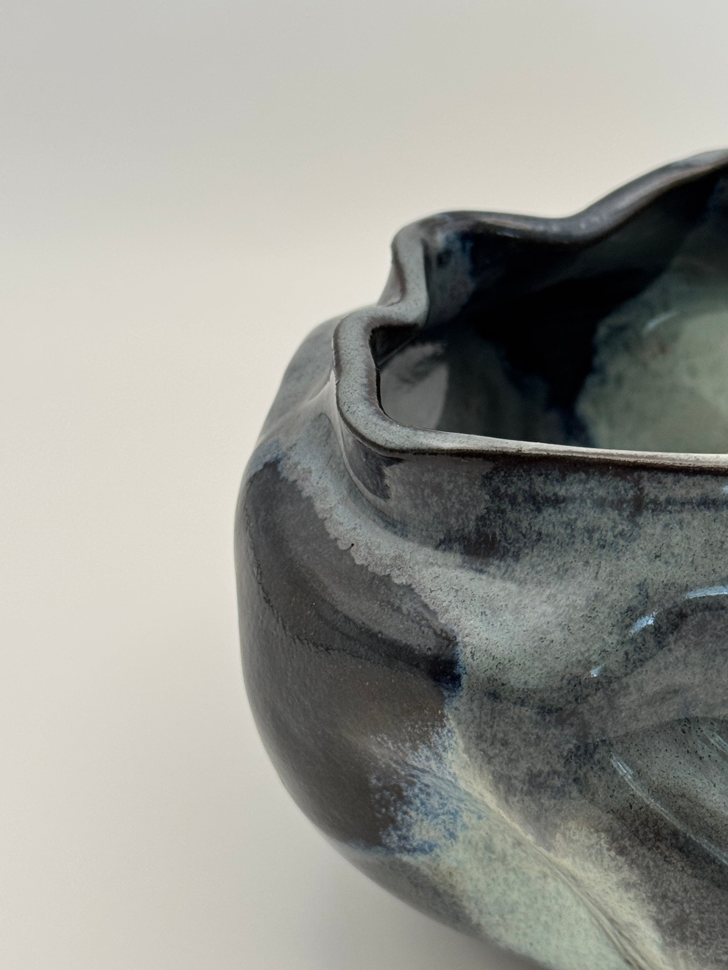 Close-up of a ceramic vessel with a dark blue and slate blue glaze, showing its textured, organic wavy rim against a neutral light gray background.