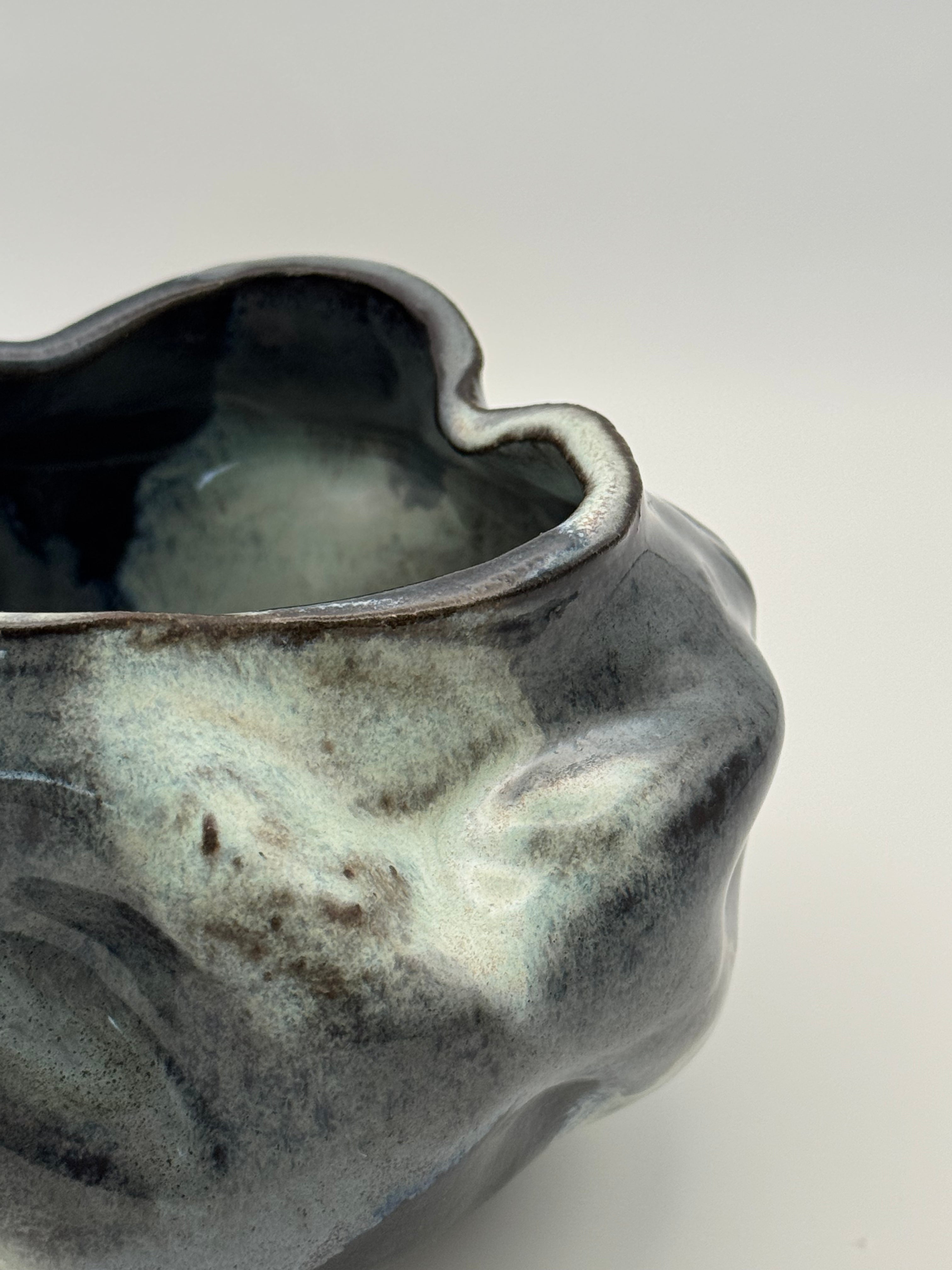 Close-up of a sculptural handmade ceramic bowl with an organic, wavy shape and a glossy, marbled glaze in darkblue, blue-gray, and light blue, set against a neutral gray background.