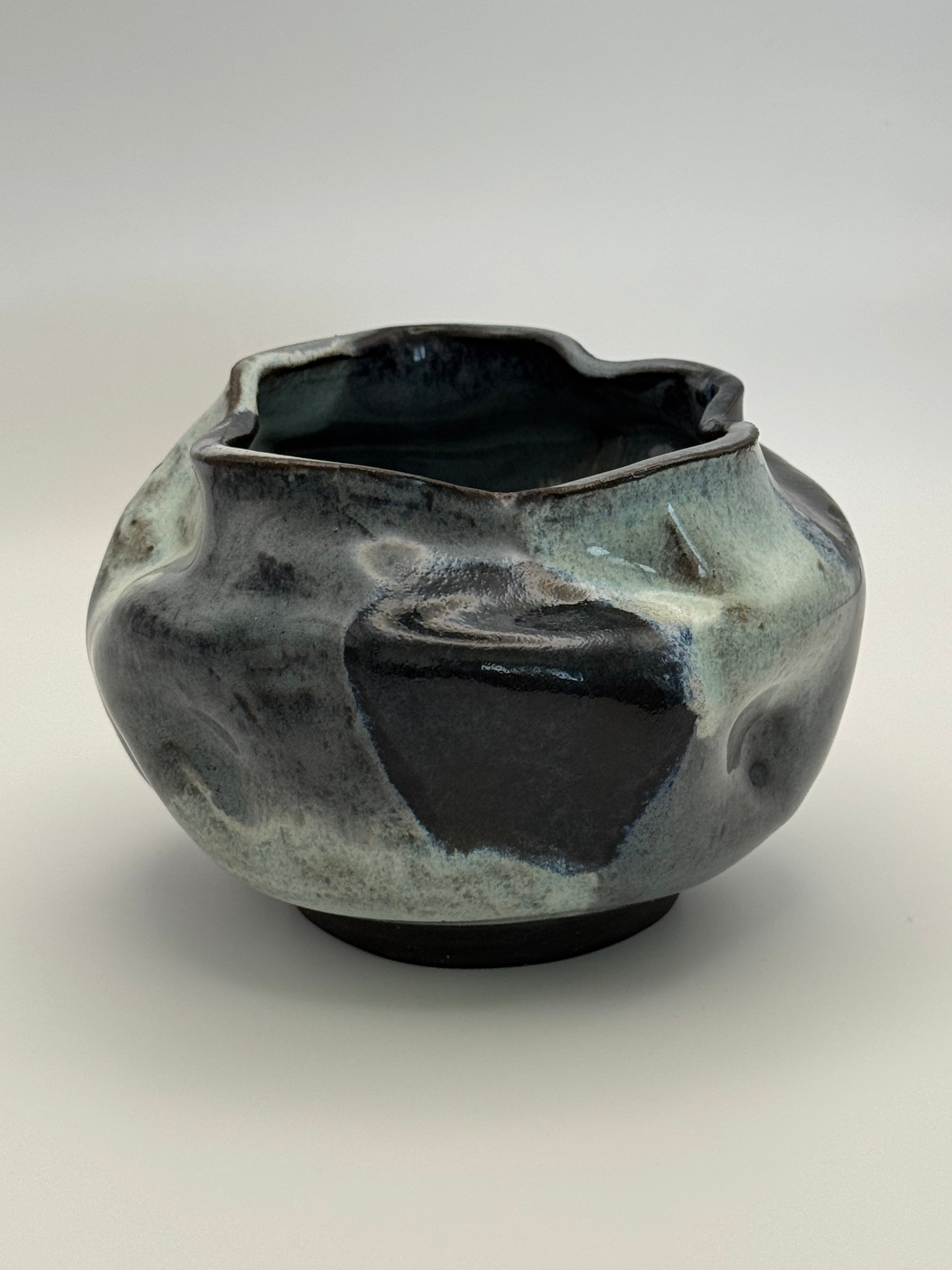 A handmade ceramic bowl with an organic, wavy shape and a glossy, marbled glaze in darkblue, blue-gray, and light blue, set against a neutral gray background.