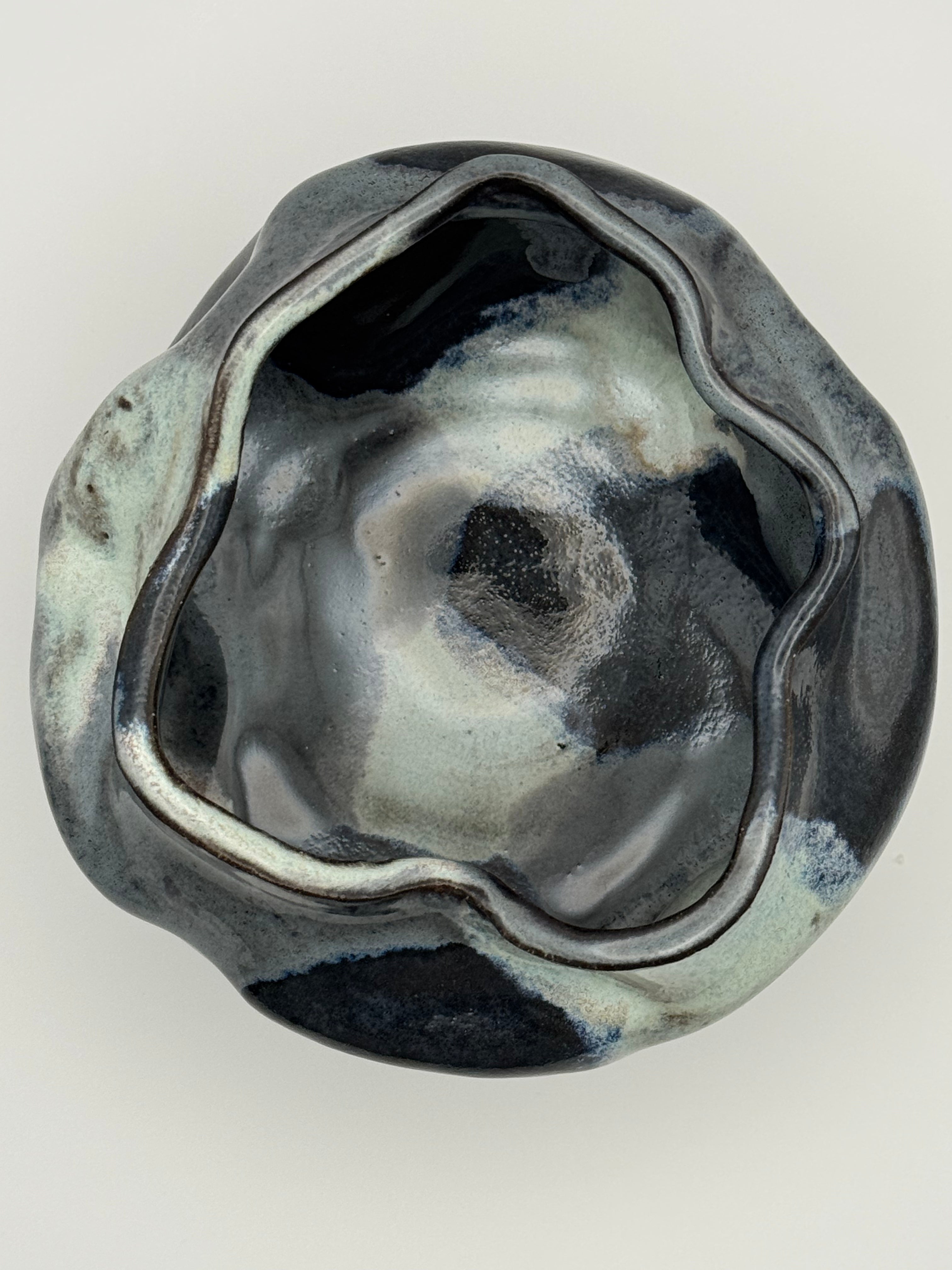 top view of A handmade ceramic bowl with an organic, wavy shape and a glossy, marbled glaze in darkblue, blue-gray, and light blue, set against a neutral gray background.