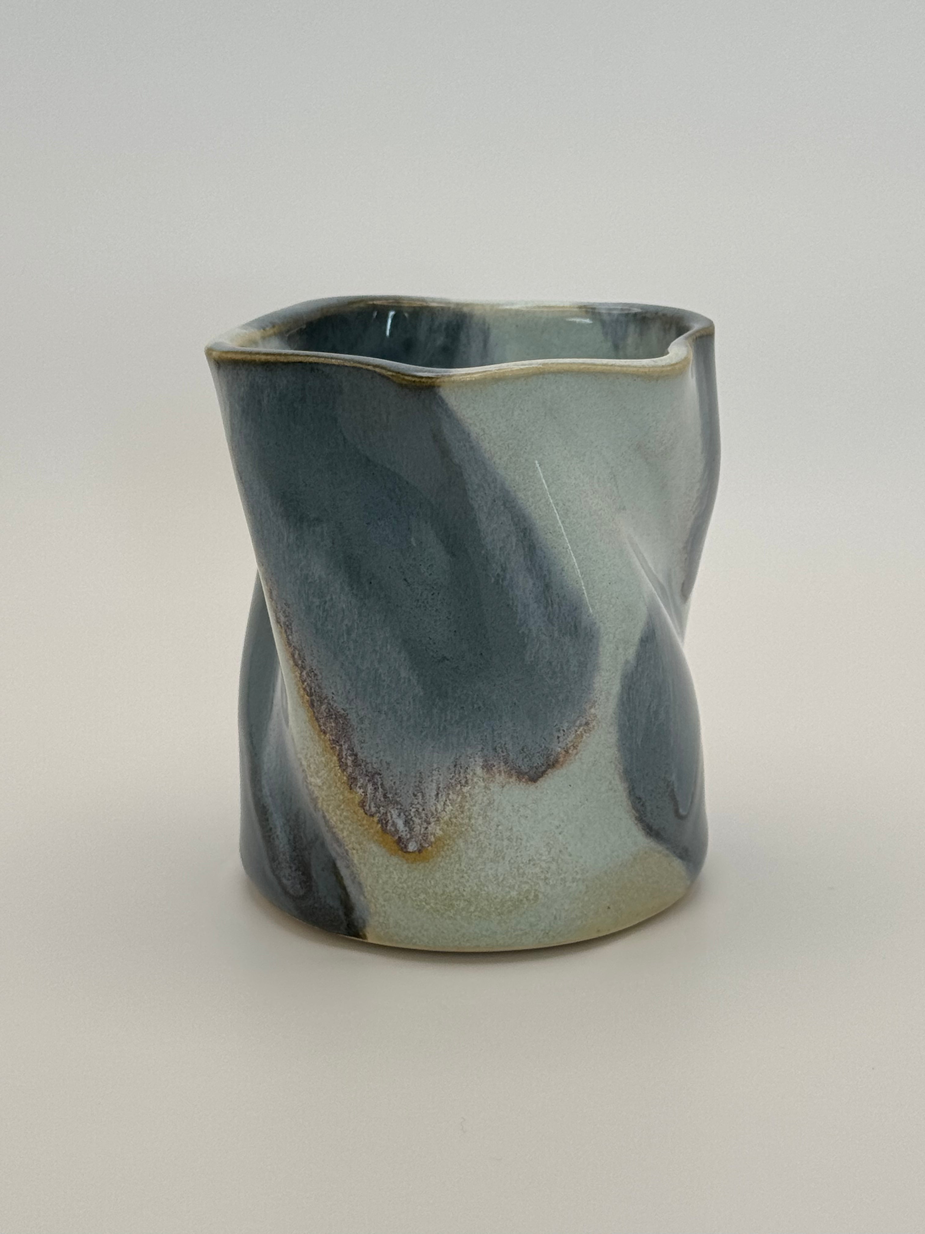 A sculptural, twisted ceramic mini vase or cup with a glossy glaze in shades of slate blue and light blue, accented with a subtle dusty rose speckles, set against a neutral light gray background.