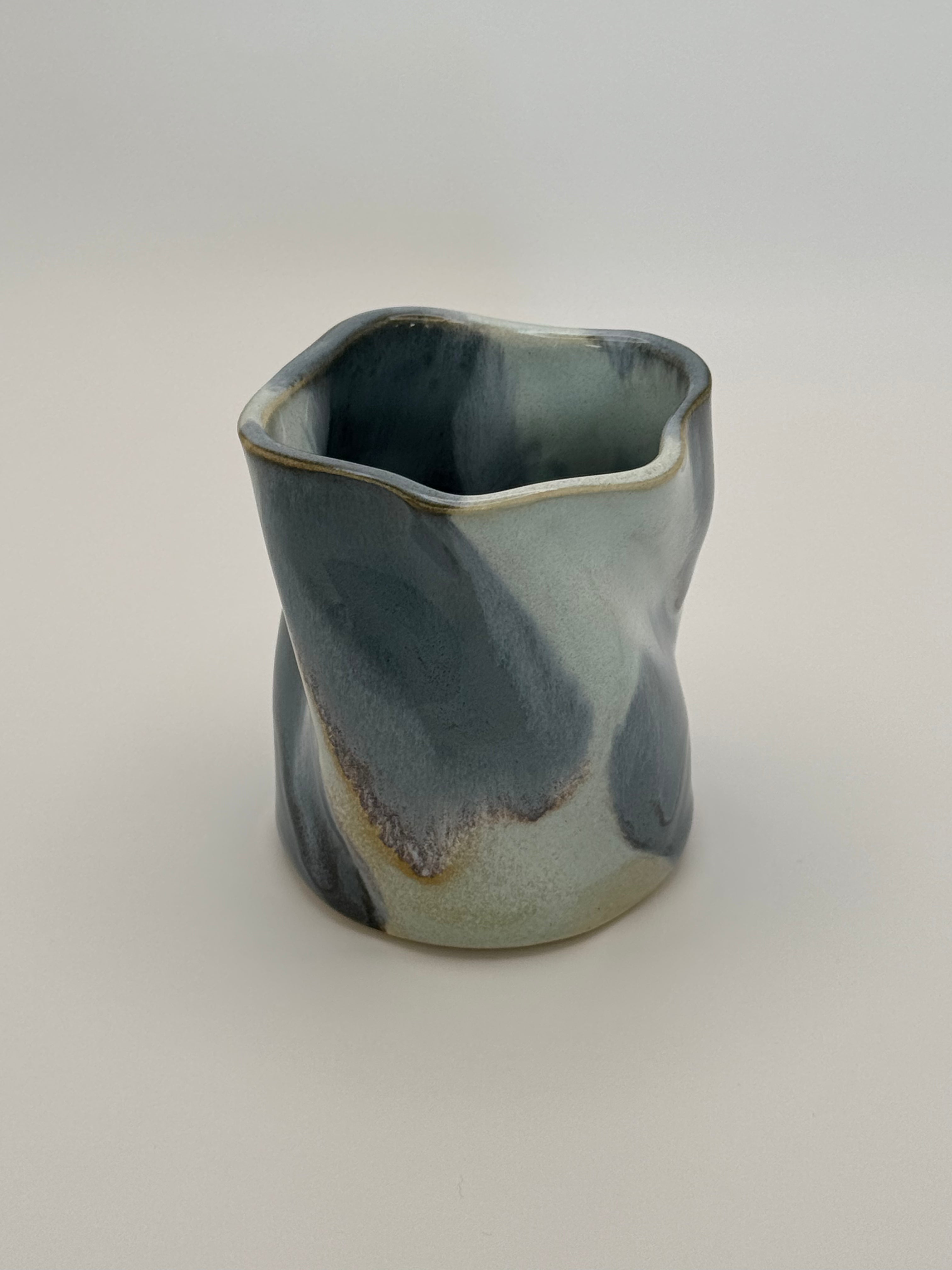 A sculptural, twisted ceramic mini vase or cup with a glossy glaze in shades of slate blue and light blue, accented with a subtle dusty rose speckles, set against a neutral light gray background.