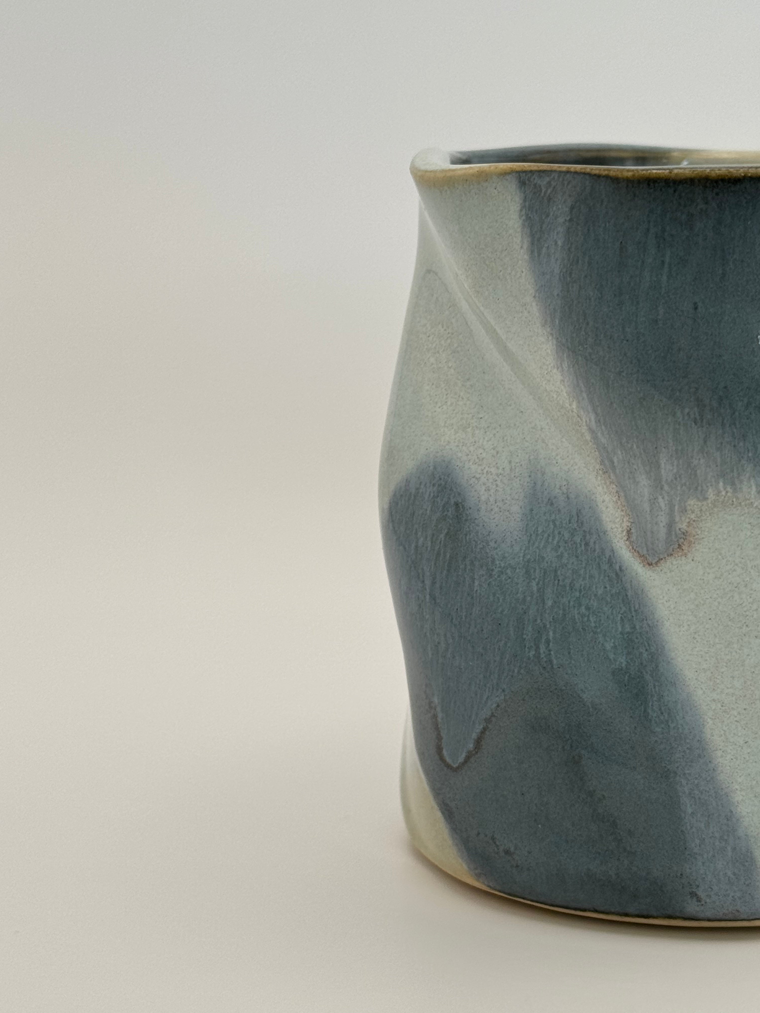 A close-up studio shot of A sculptural, twisted ceramic mini vase or cup with a glossy glaze in shades of slate blue and light blue, accented with a subtle dusty rose speckles, set against a neutral light gray background.