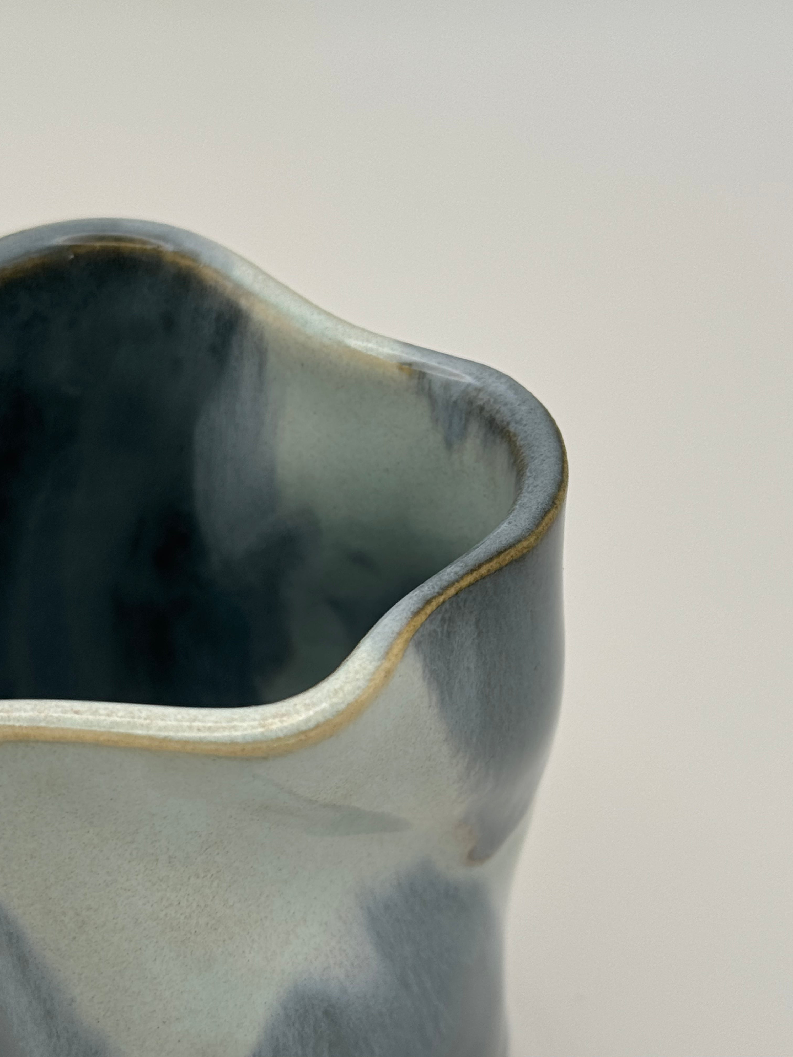 close up of the rom of A sculptural, twisted ceramic mini vase or cup with a glossy glaze in shades of slate blue and light blue, accented with a subtle dusty rose speckles, set against a neutral light gray background.