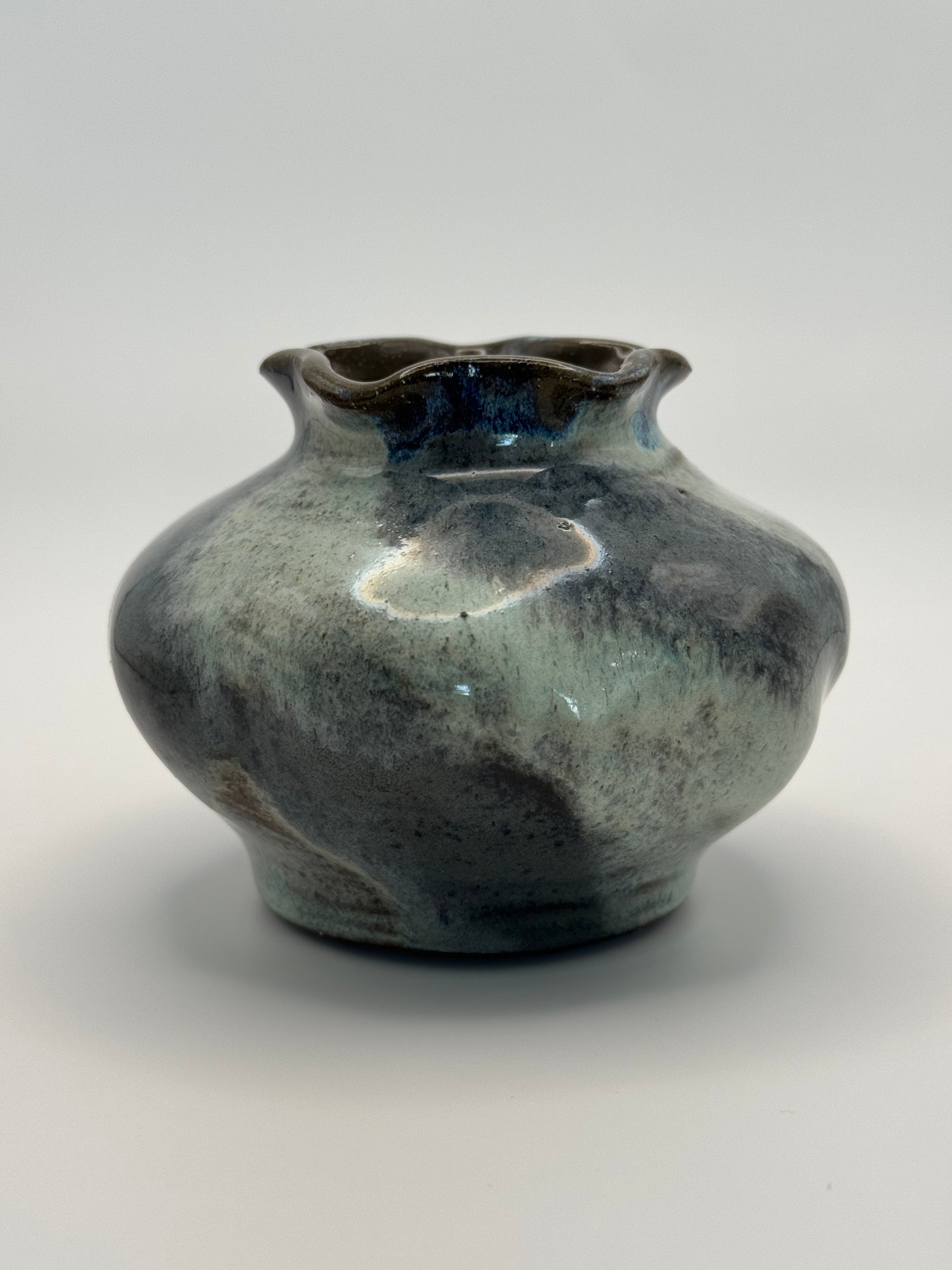 A handmade ceramic bud vase with a rounded, bulbous shape and a flared, scalloped rim. It features a mottled glaze with a blend of blue-gray and deep blue with a glossy highlight reflecting light.