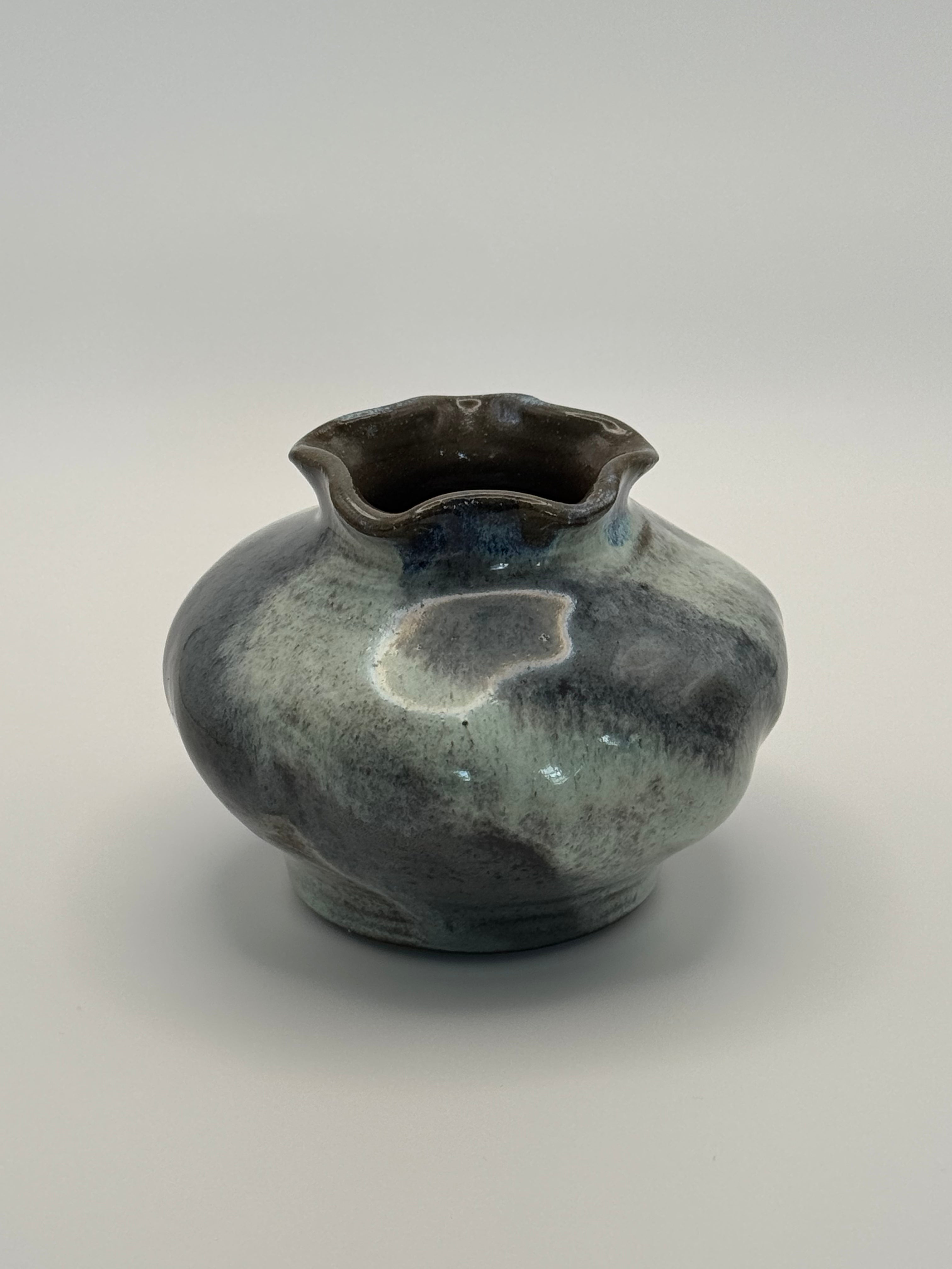 A handmade ceramic vase with a rounded body, flared scalloped rim, and a textured glaze featuring a mix of dark blue and light blue-grey with a glossy finish, set against a neutral gray background.