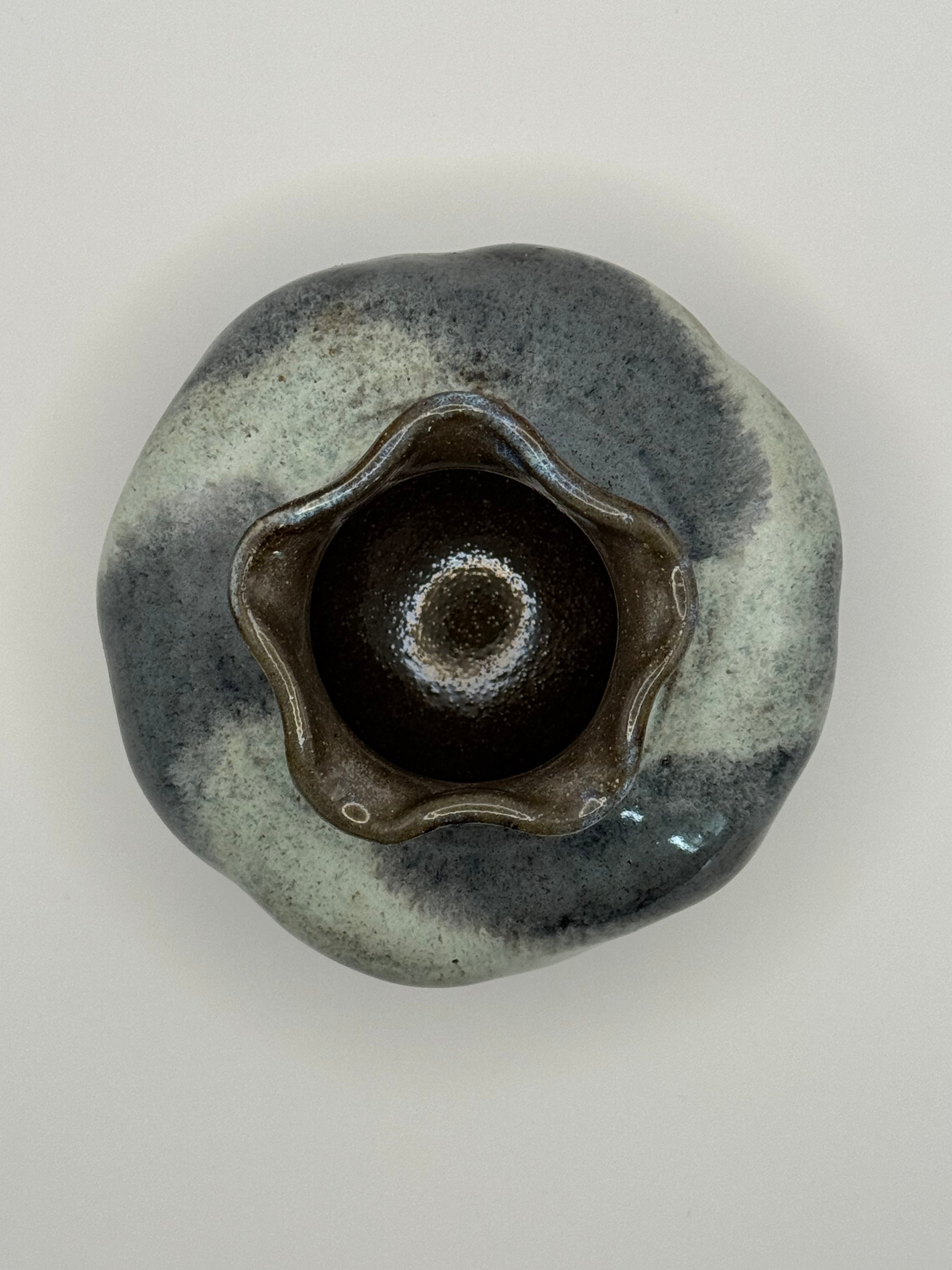 A handmade ceramic bud vase with a dark brown interior and a mottled exterior of blue-grey and dark blue glaze, photographed from directly above on a light gray background.