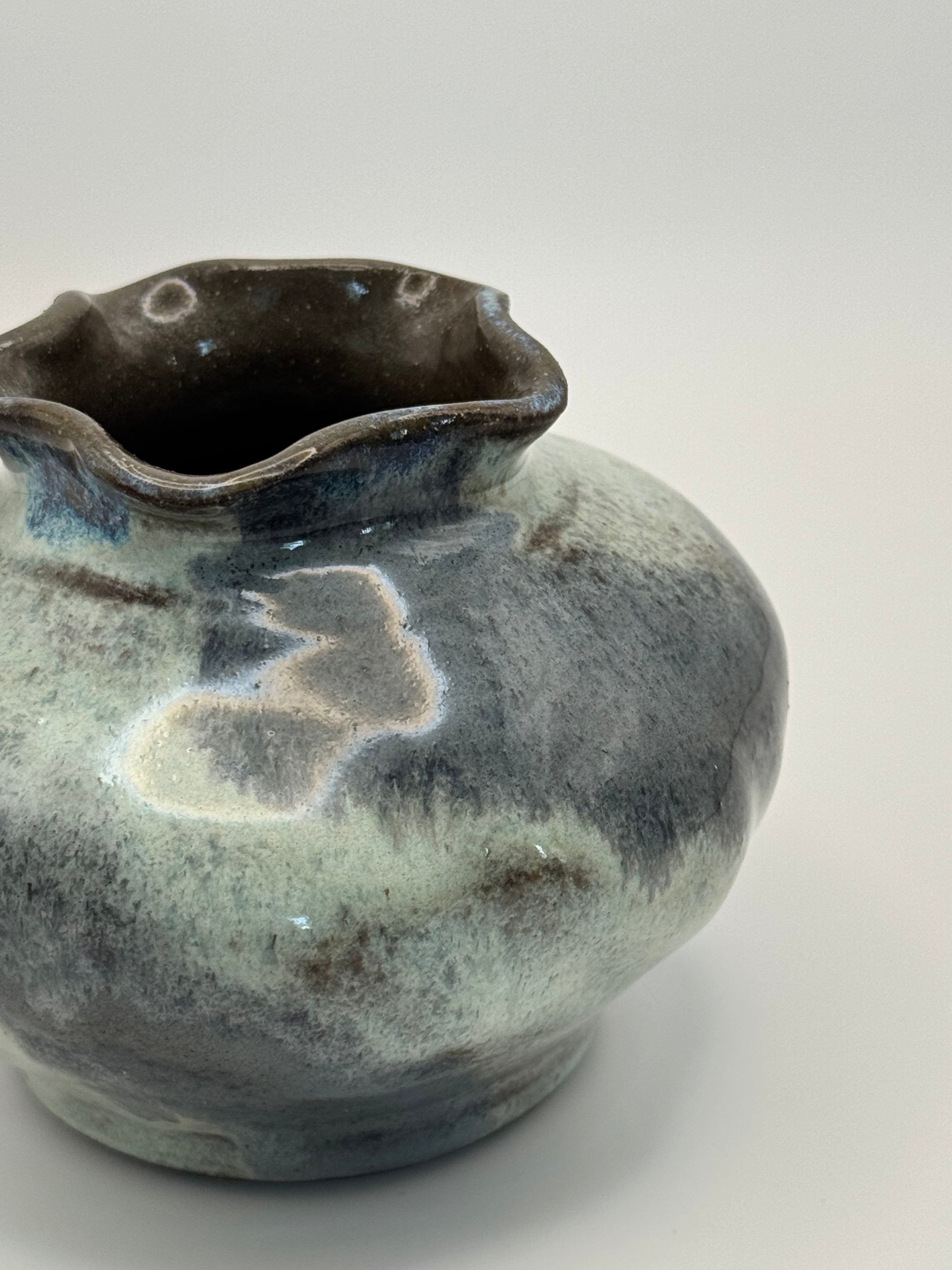 A handmade ceramic pitcher with a rounded body and a wavy, scalloped rim, featuring a mottled glaze of blue grey and dark blue with clear glaze on the interior on a light gray background.