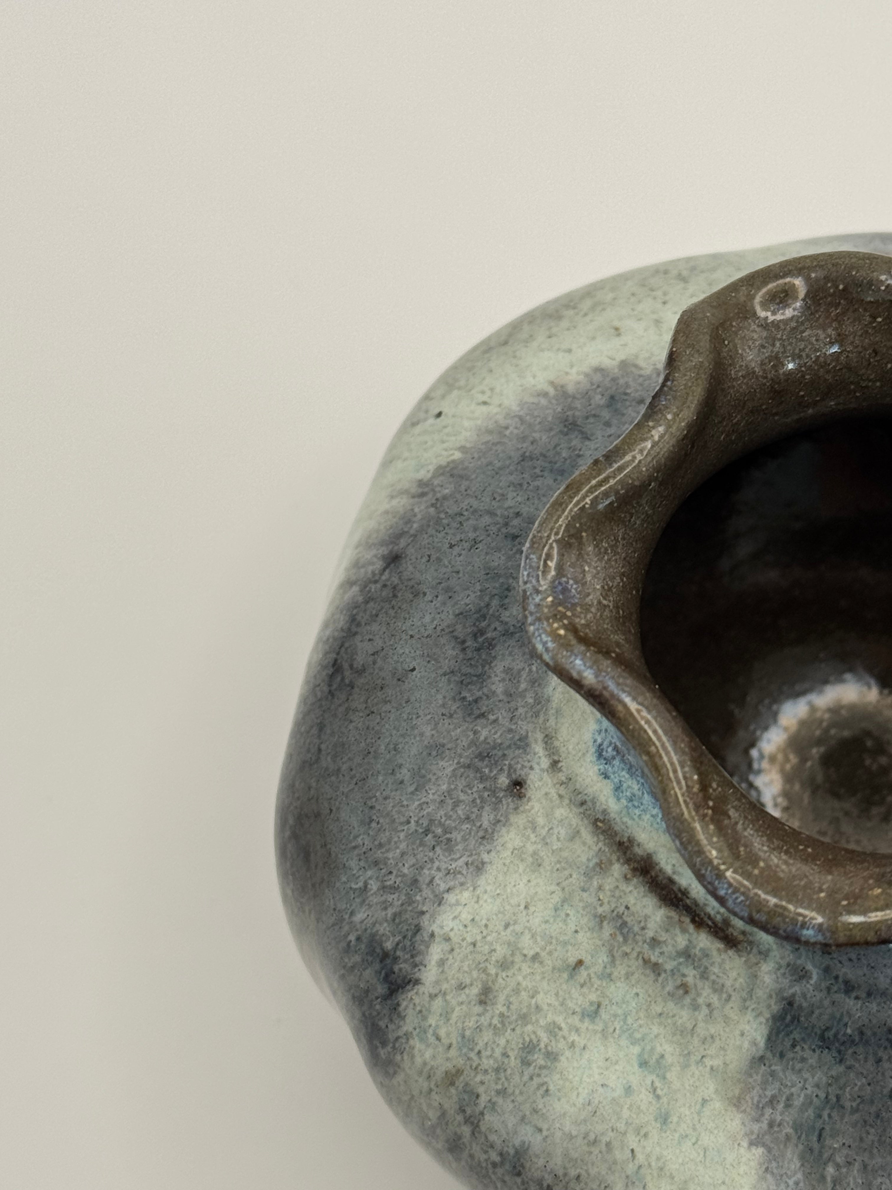 Close-up of a ceramic vessel showing its textured glaze with mottled dark blue, blue-grey, and dark brown tones of the clay, set against a plain light background.
