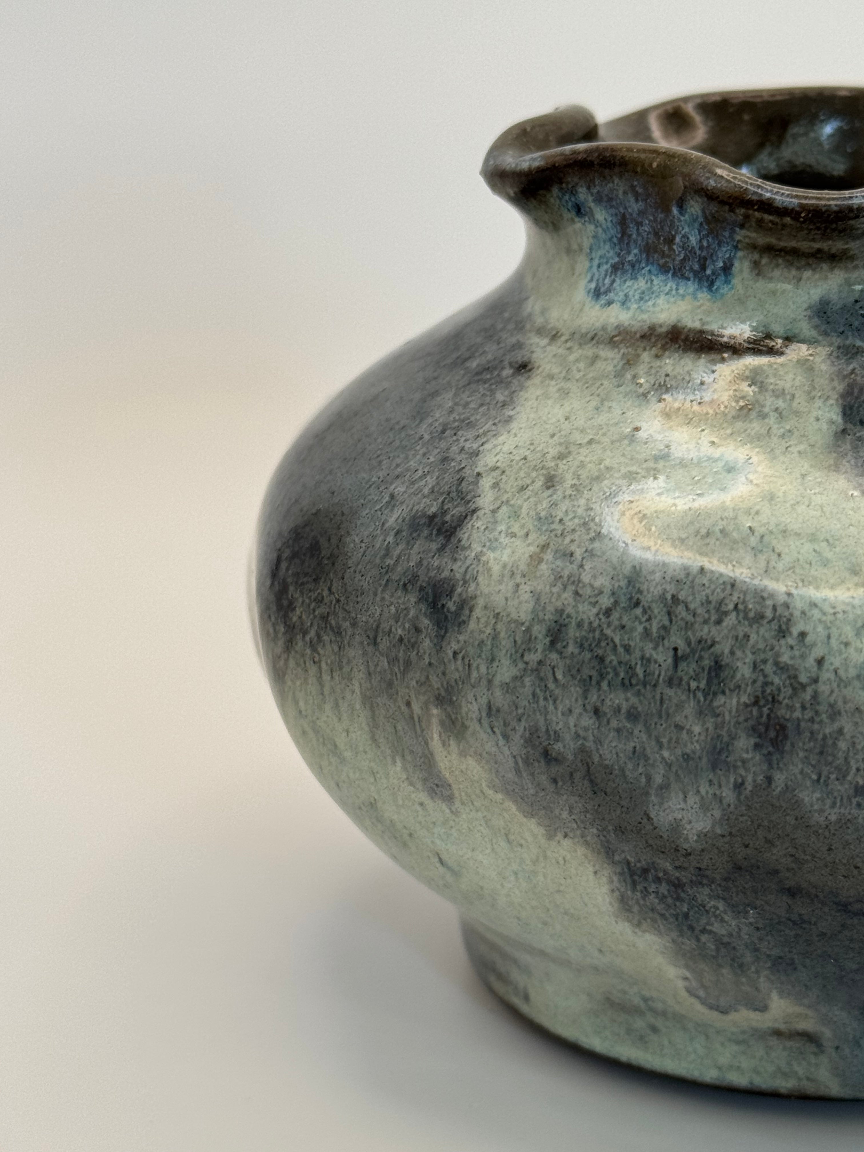 Close-up of a ceramic vase with a rounded body and flared rim, featuring a mottled glaze in dark blue and muted blue-grey on a light neutral background.