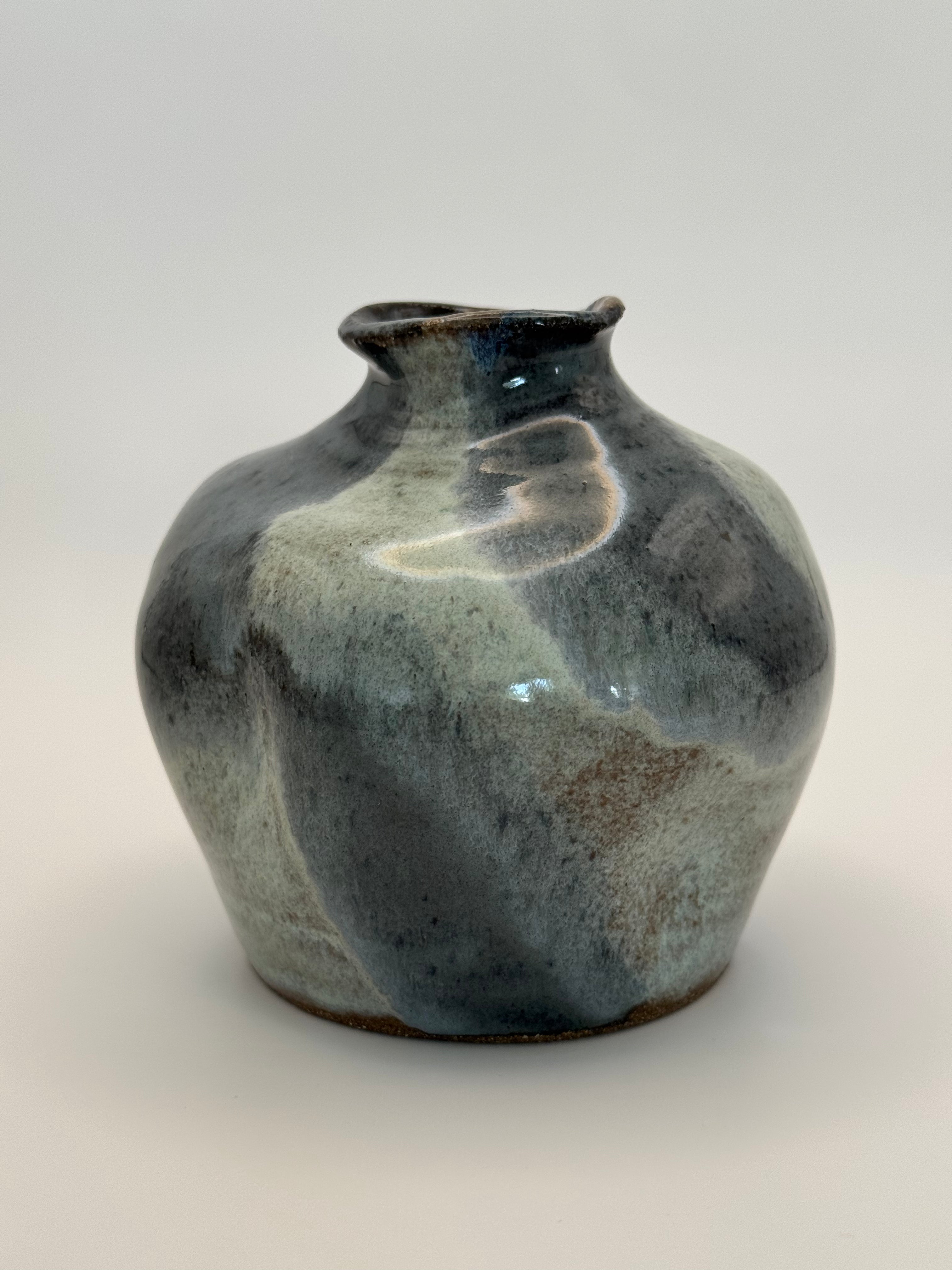 A handmade ceramic vase with a rounded body and small flared rim, featuring a mottled glaze in shades of slate blue, gray, and brown with a glossy finish, set against a plain light gray background.