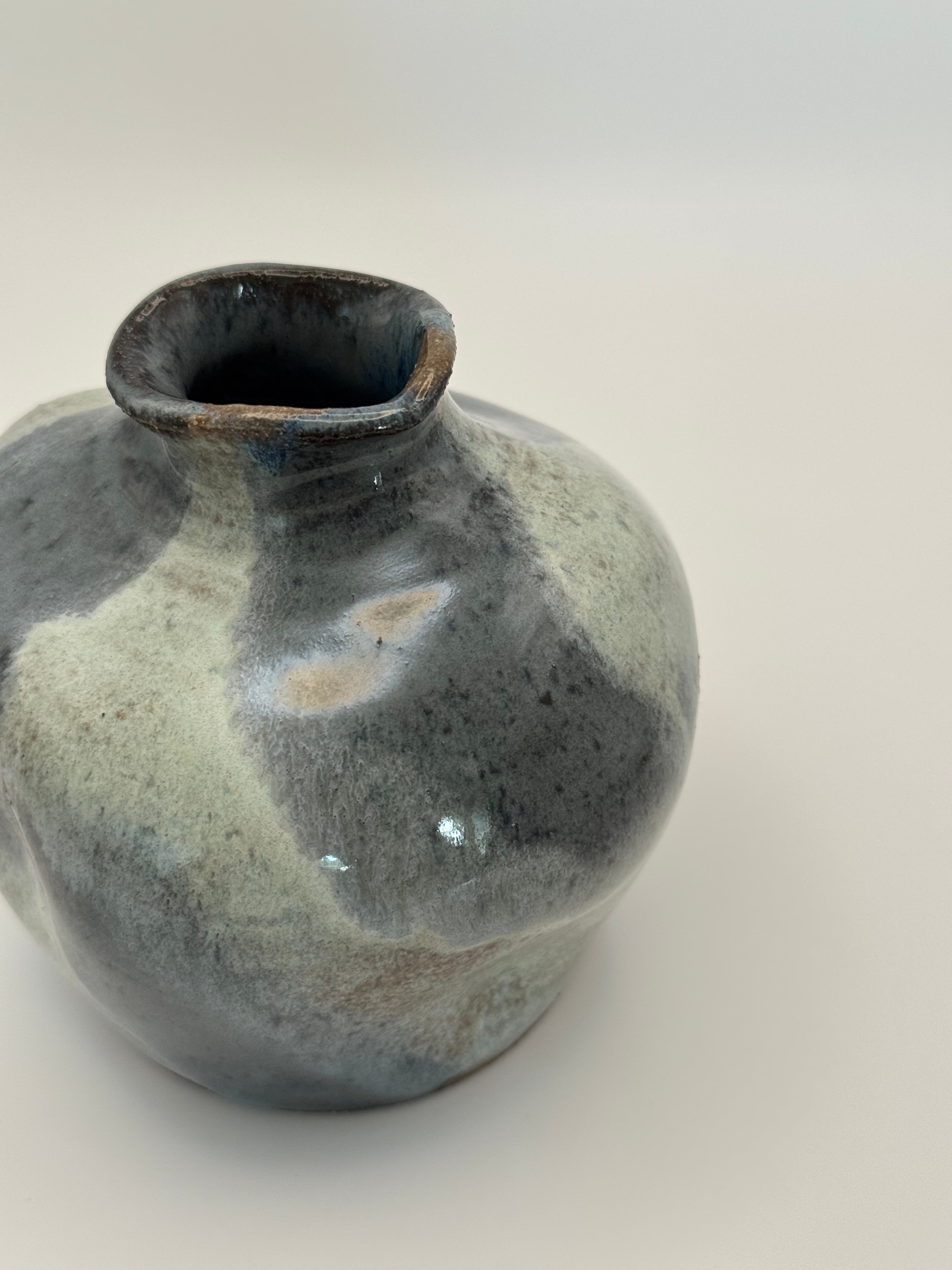 A handmade ceramic vase with a rounded bulbous body and a small flared rim, featuring a mottled glaze in shades of slate blue and blue-gray on a light neutral background.