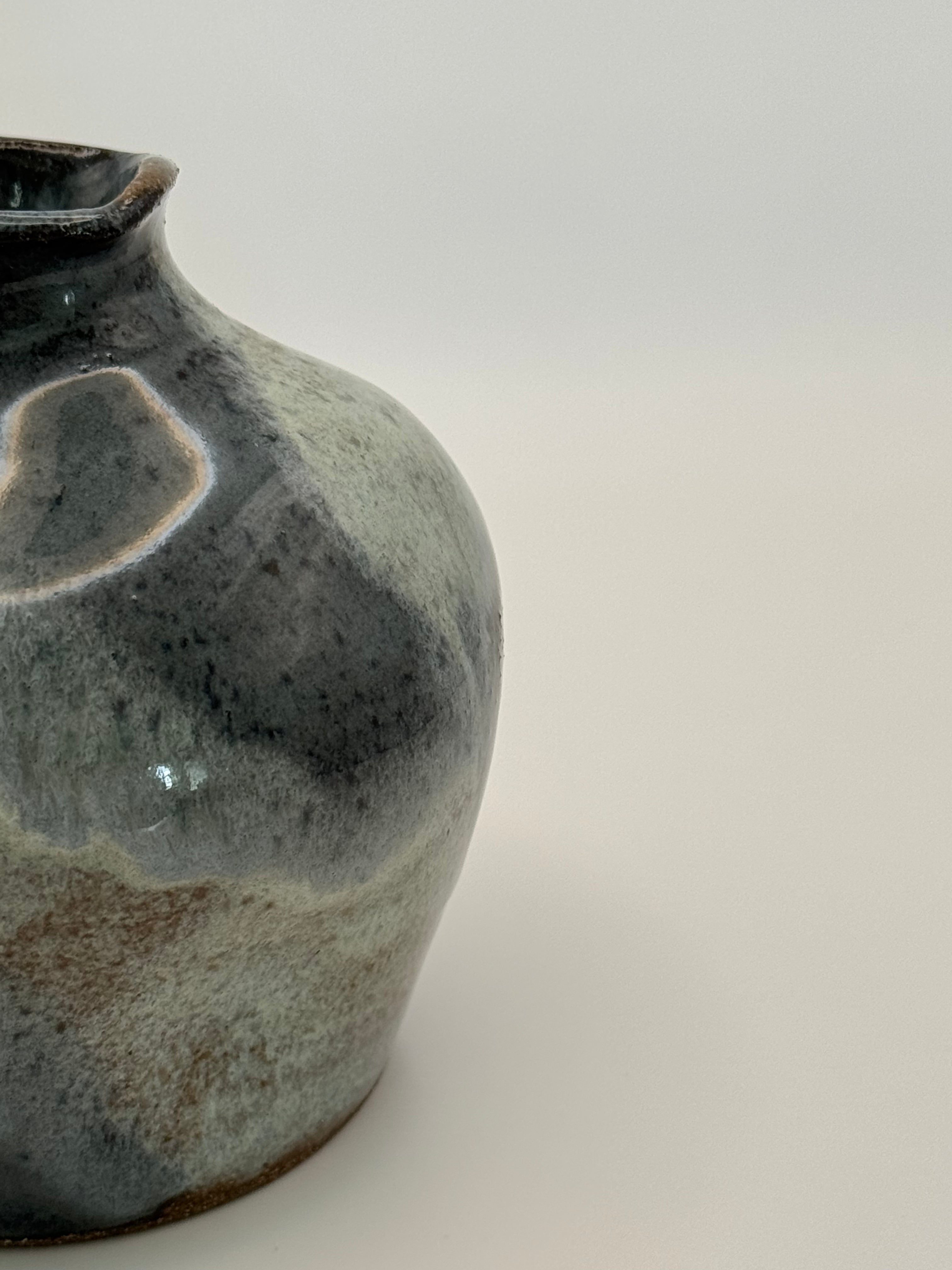 Close-up of a ceramic vase with a rounded body and short neck, featuring a mottled glaze in dark blue, blue-grey, and warm brown clay showing through the glaze on a light gray background.