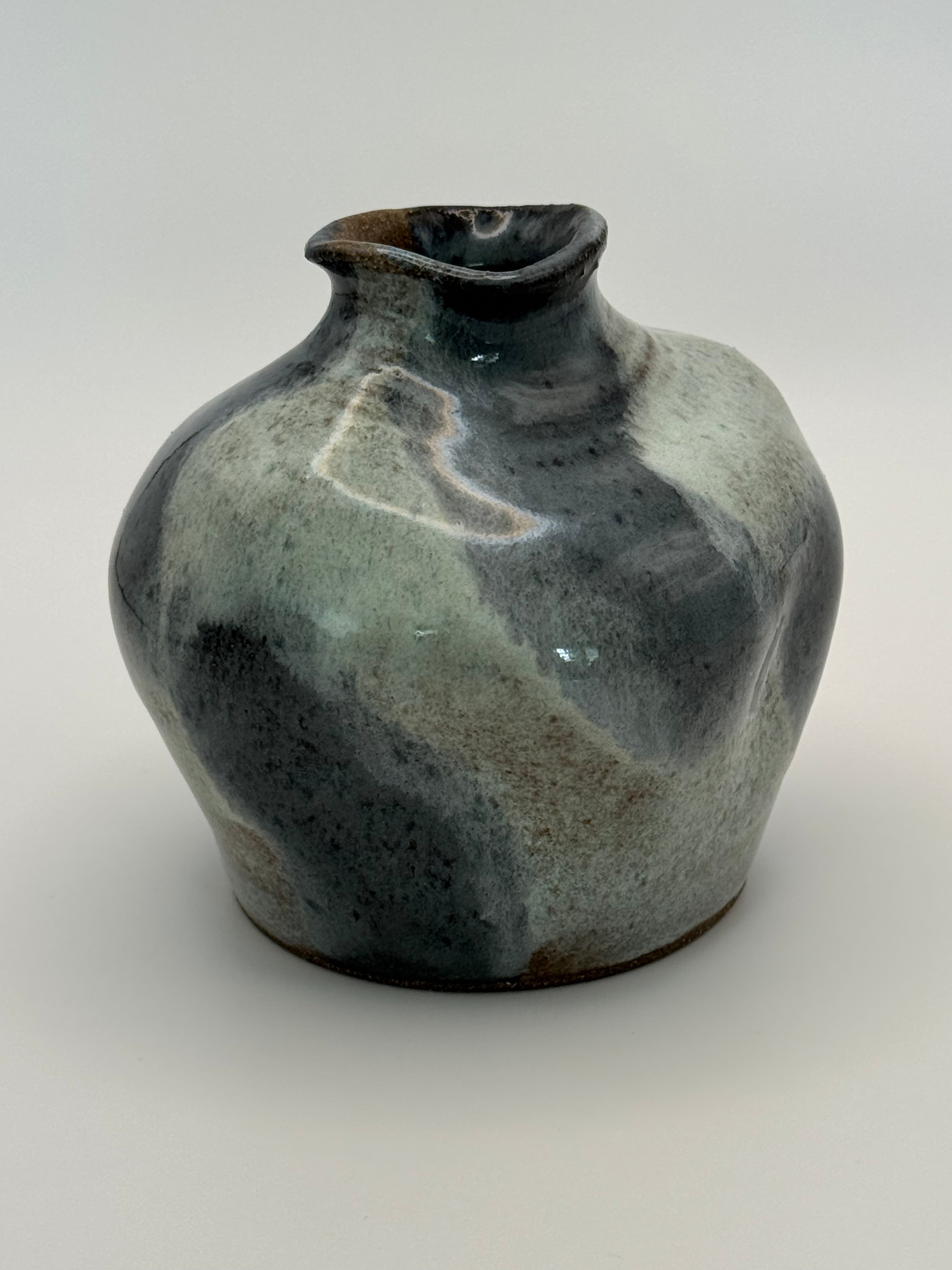 A handmade ceramic vase with a rounded body, small flared rim, and glossy glaze featuring mottled shades of blue-grey, dark blue, and muted blue on a light gray background.