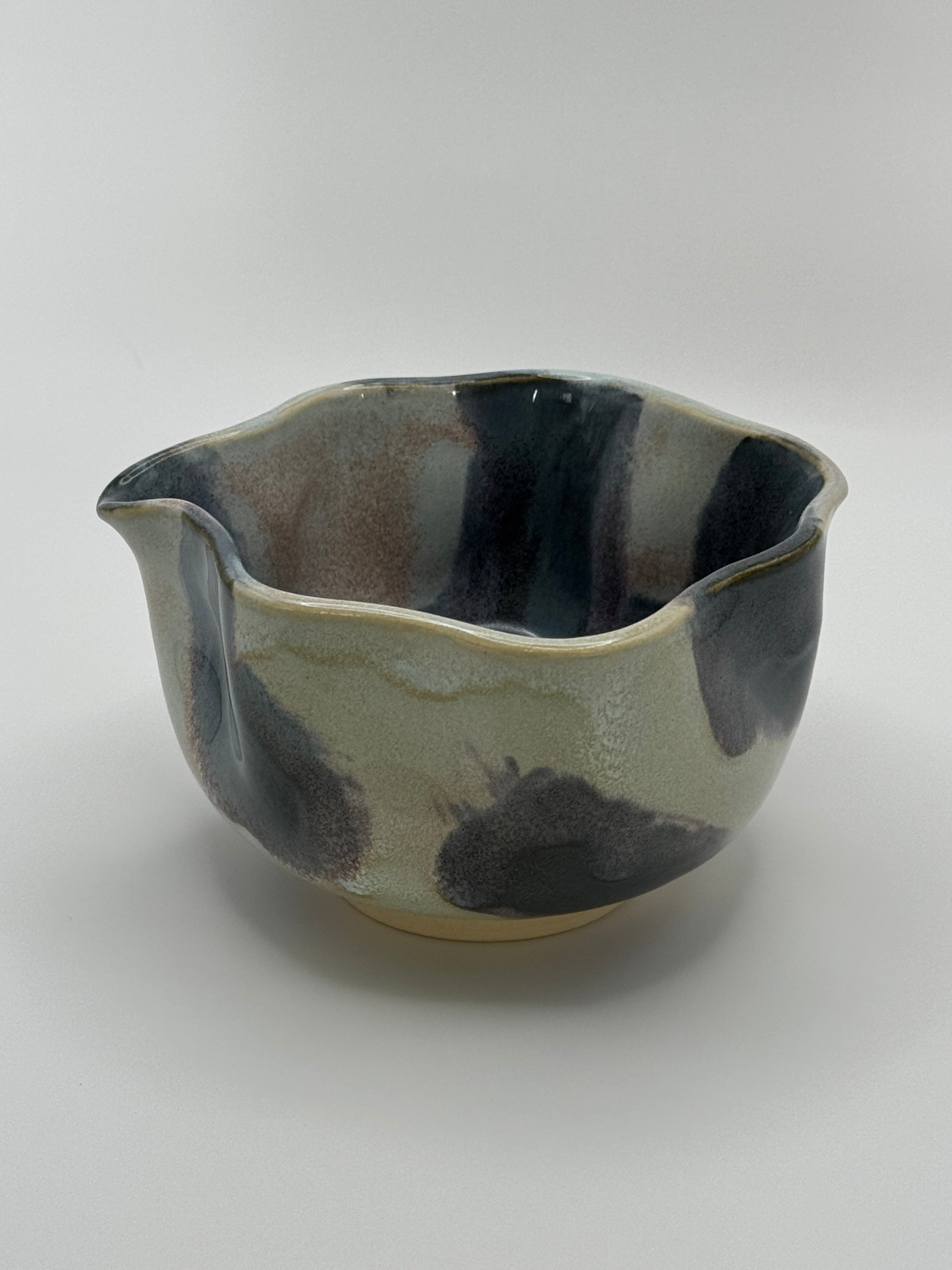 A handmade ceramic bowl with a wavy rim and a small footed base, featuring a glossy glaze with abstract slated blue and light blue brushstrokes over a white clay base, set against a plain light gray background.