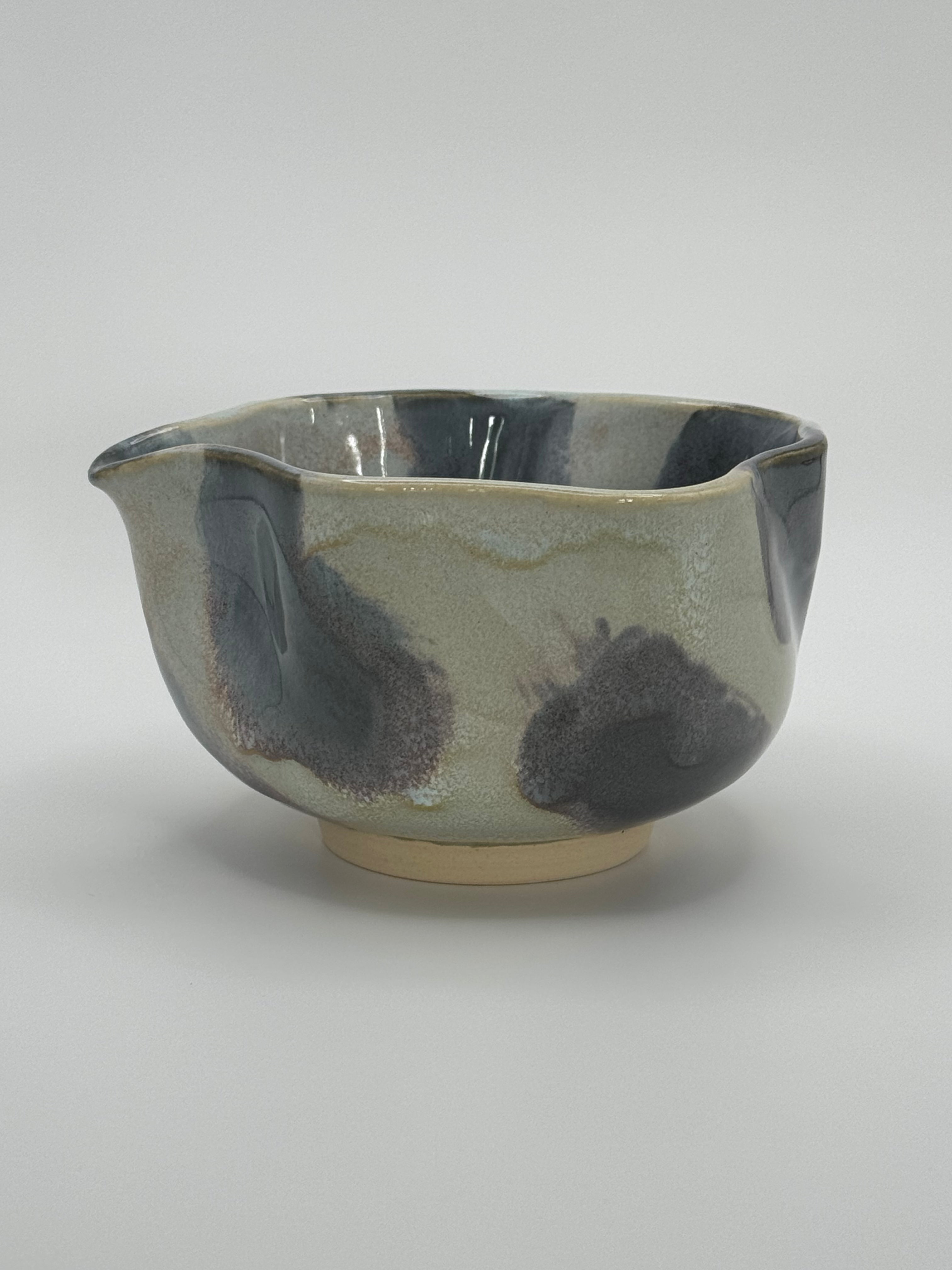 A handmade ceramic bowl with a wavy rim and a small footed base, featuring a glossy glaze with abstract slated blue and light blue brushstrokes over a white clay base, set against a plain light gray background.