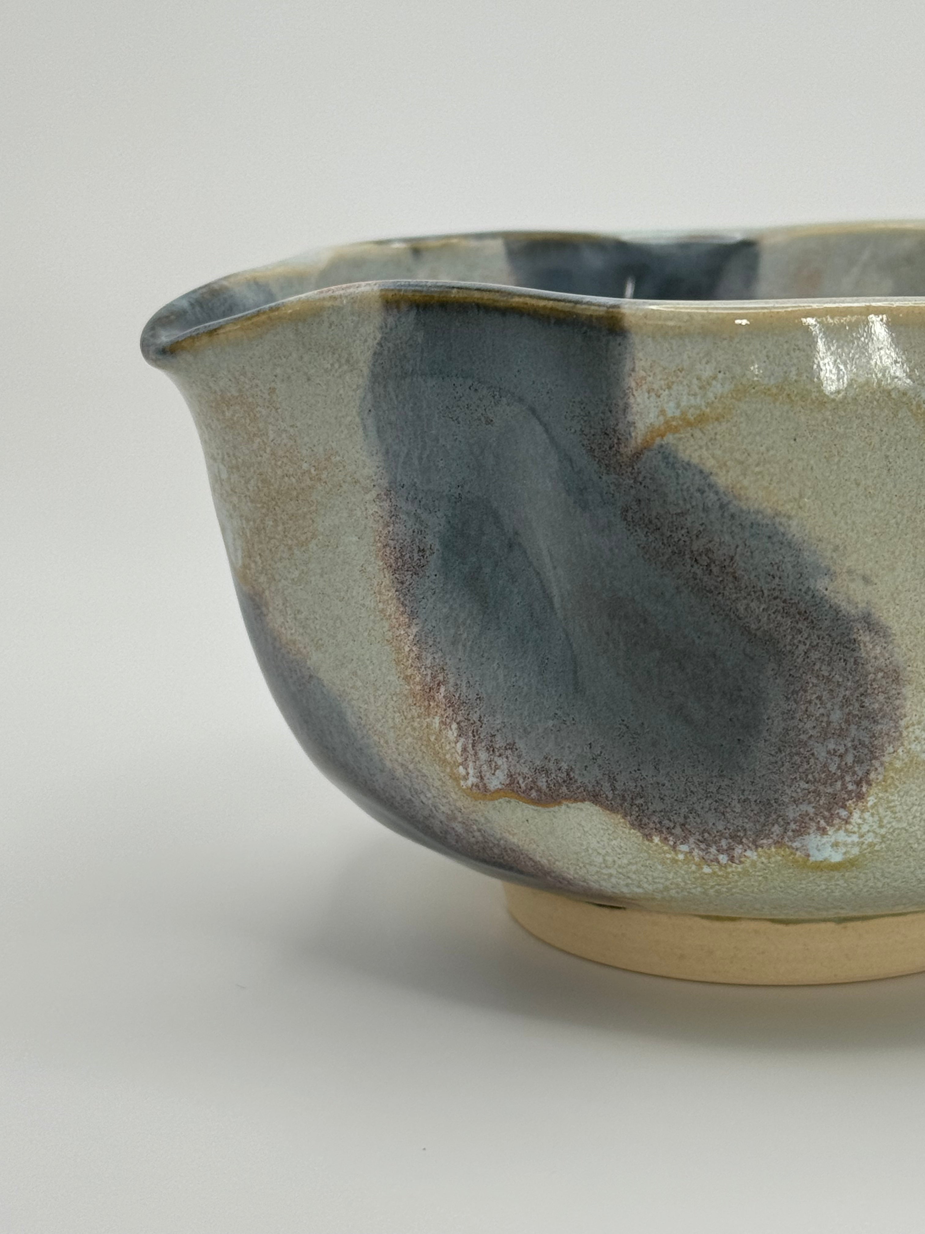 A close-up of ceramic bowl with a wavy rim and a small footed base, featuring a glossy glaze with abstract slated blue and light blue brushstrokes over a white clay base, set against a plain light gray background.