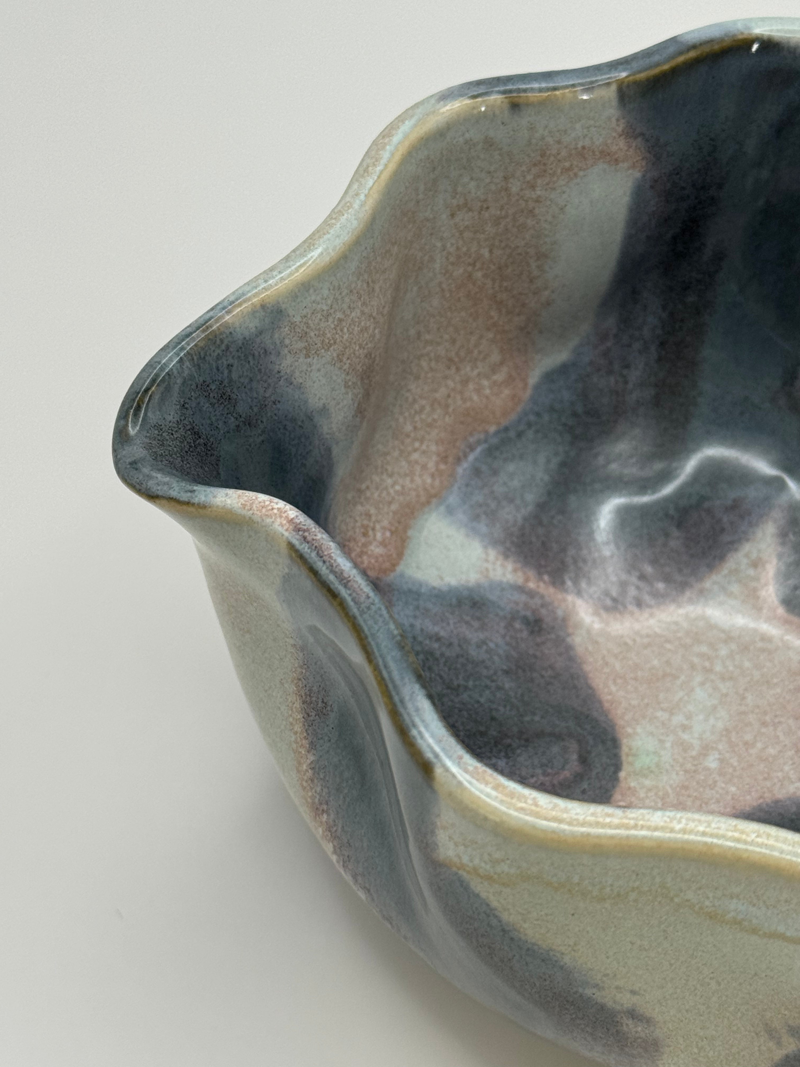 Close-up of a ceramic bowl with a wavy, scalloped rim, featuring a marbled glaze in shades of slate blue, dusty rose specks, and light blue  in the interior on a light gray background.