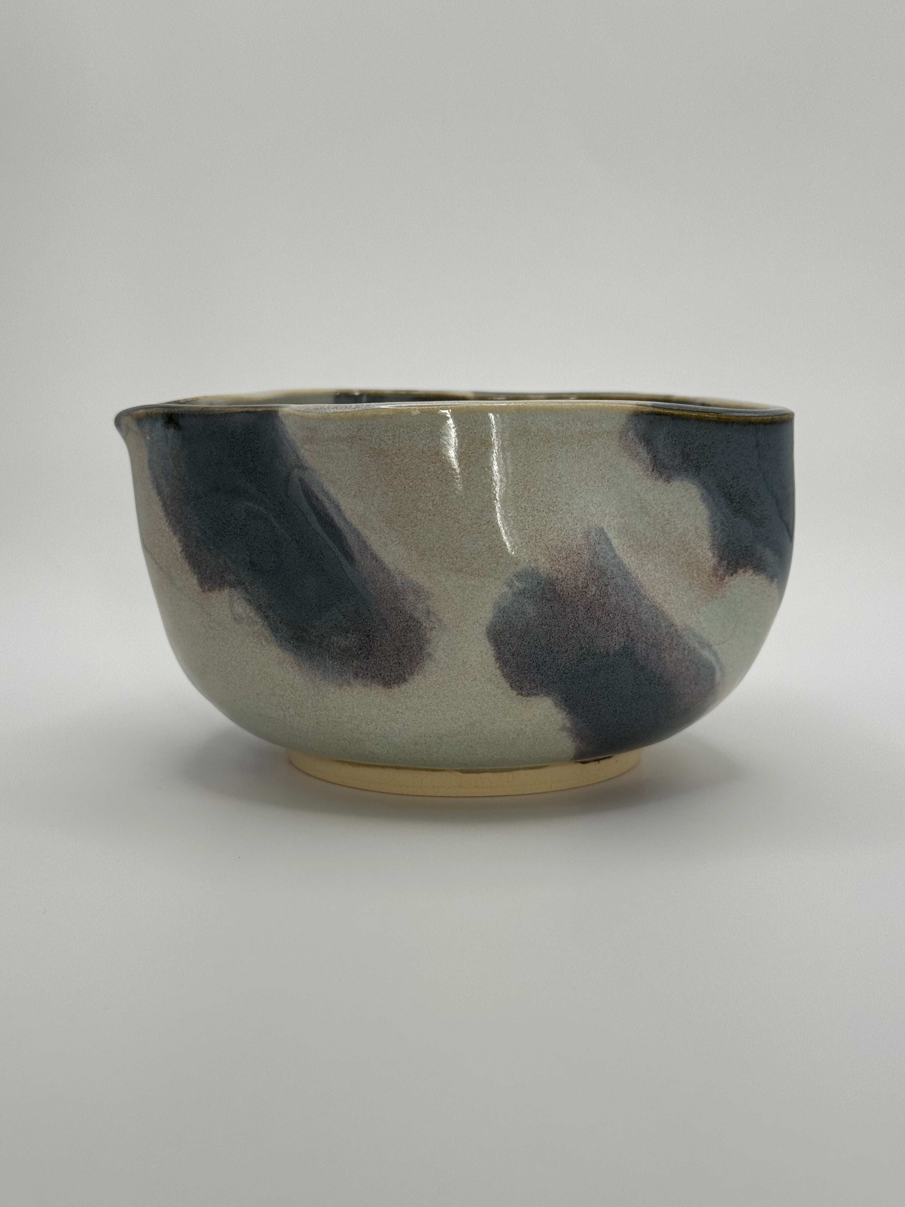 A handmade ceramic bowl with a wavy rim and a small footed base, featuring a glossy glaze with abstract slated blue and light blue brushstrokes over a white clay base, set against a plain light gray background.
