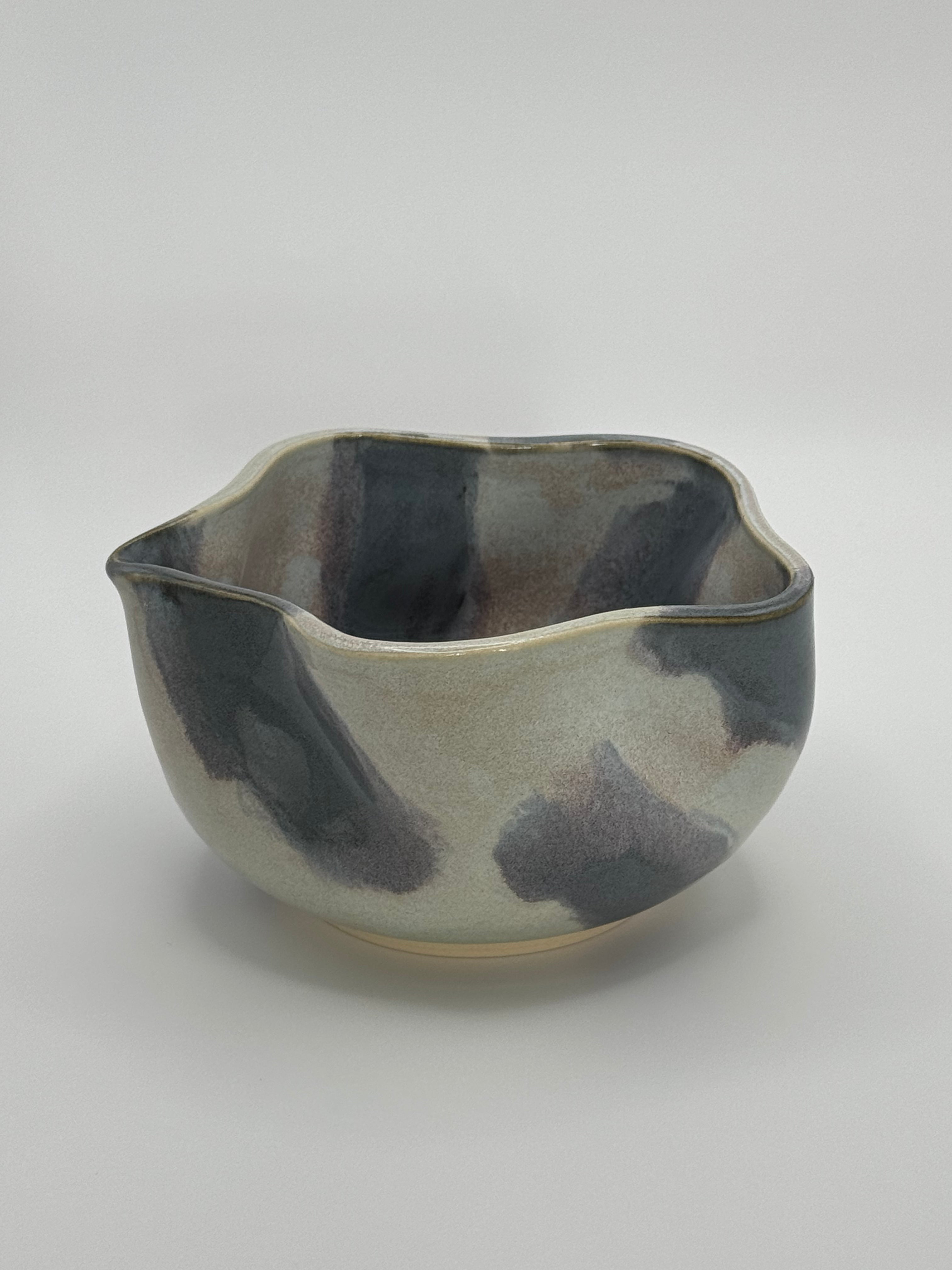A handmade ceramic bowl with a wavy rim and a small footed base, featuring a glossy glaze with abstract slated blue and light blue brushstrokes over a white clay base, set against a plain light gray background.