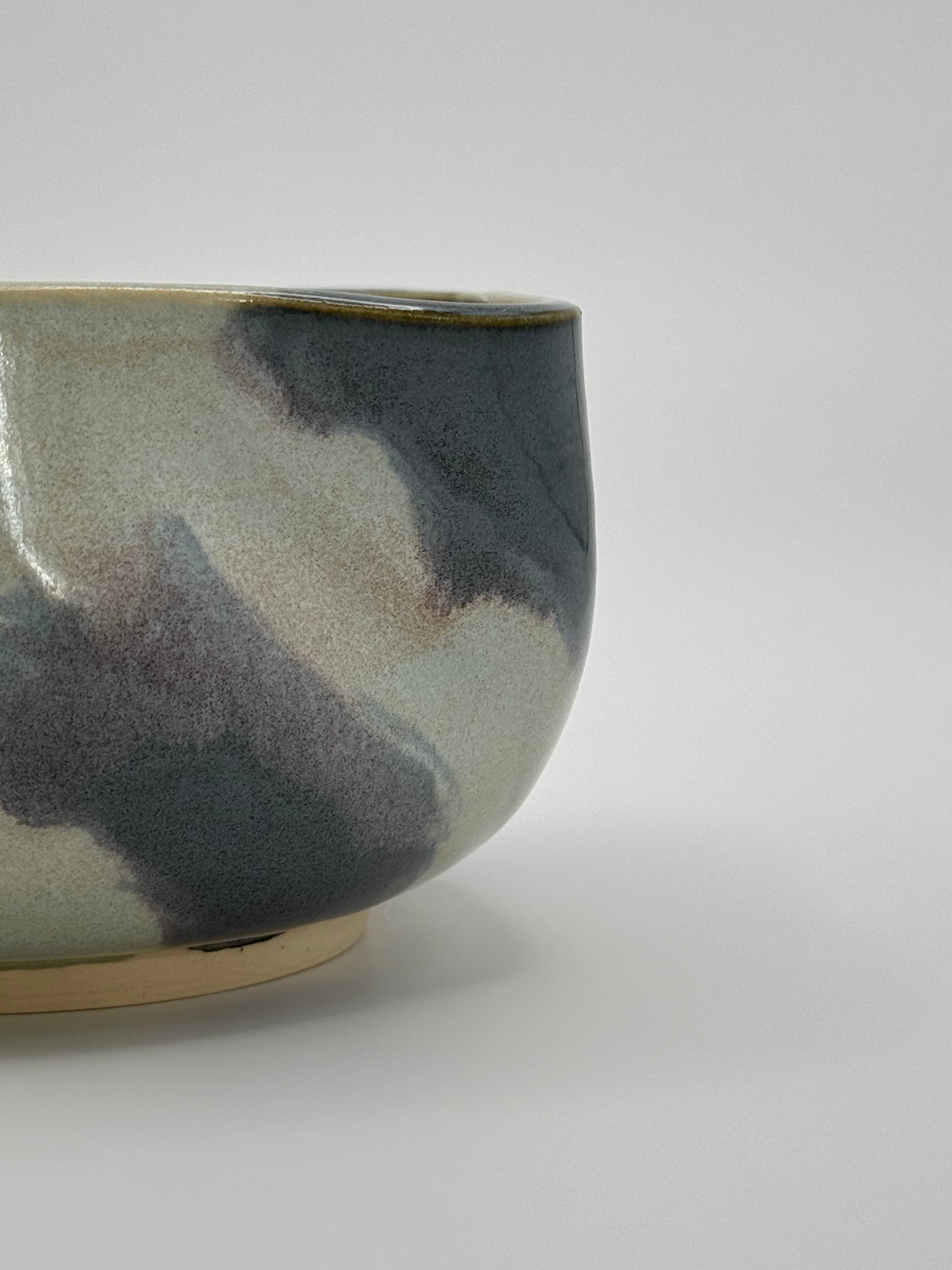 A close-up of A handmade ceramic bowl with a wavy rim and a small footed base, featuring a glossy glaze with abstract slated blue and light blue brushstrokes over a white clay base, set against a plain light gray background.
