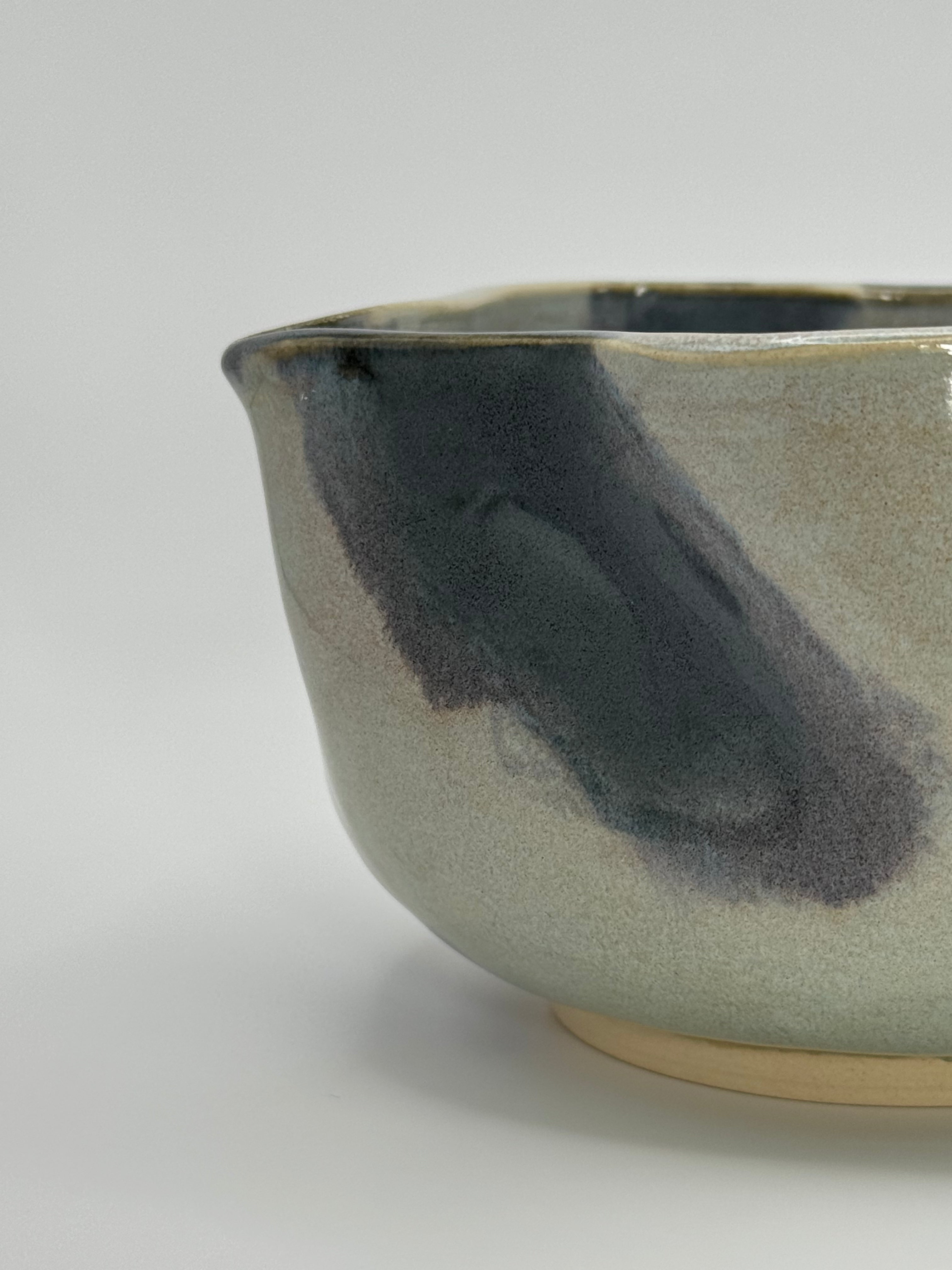 Close-up of A handmade ceramic bowl with a wavy rim and a small footed base, featuring a glossy glaze with abstract slated blue and light blue brushstrokes over a white clay base, set against a plain light gray background.