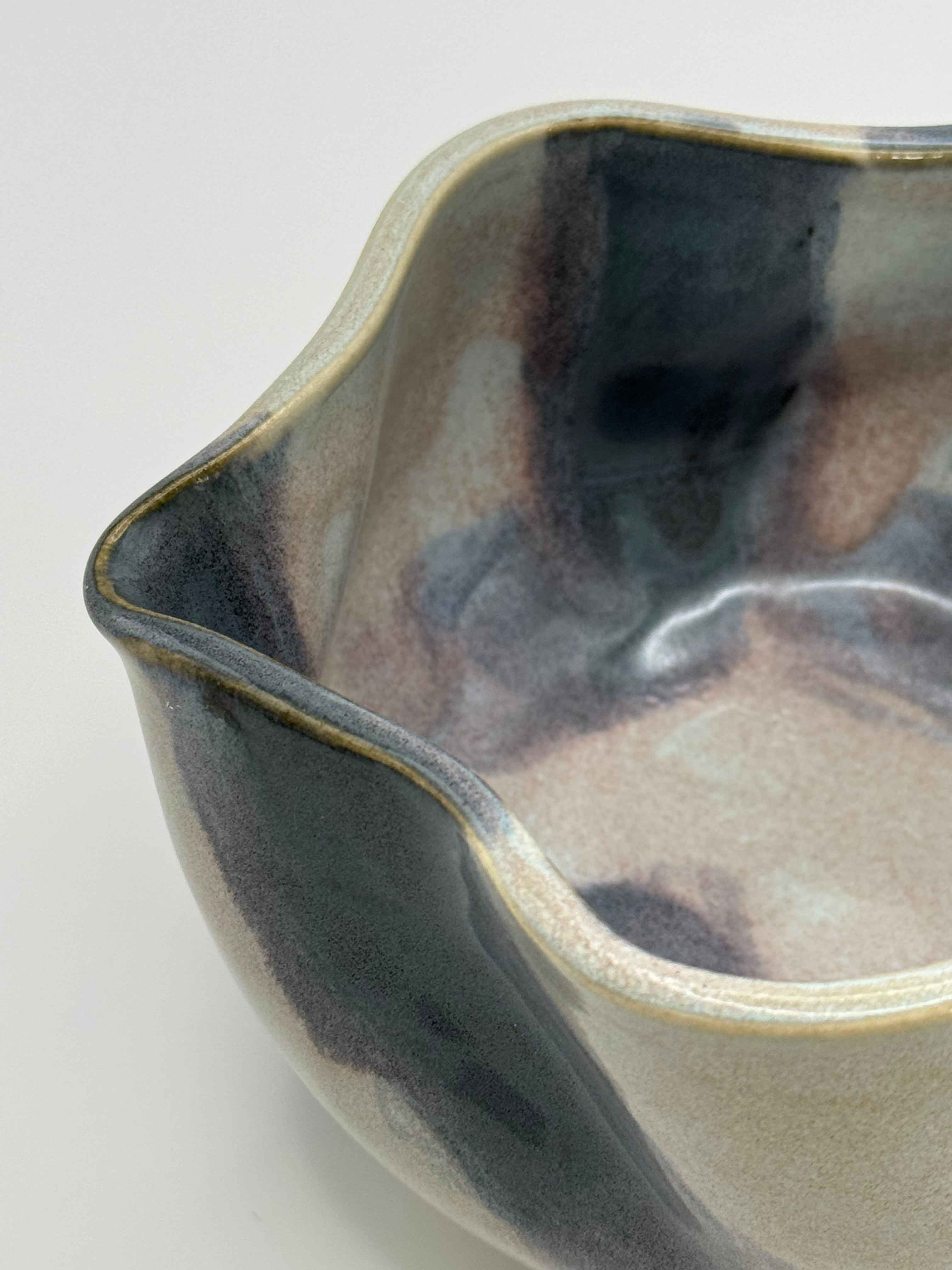 Close-up of A handmade ceramic bowl with a wavy rim and a small footed base, featuring a glossy glaze with abstract slated blue and light blue brushstrokes over a white clay base with dusty rose specks in the interior, set against a plain light gray background.
