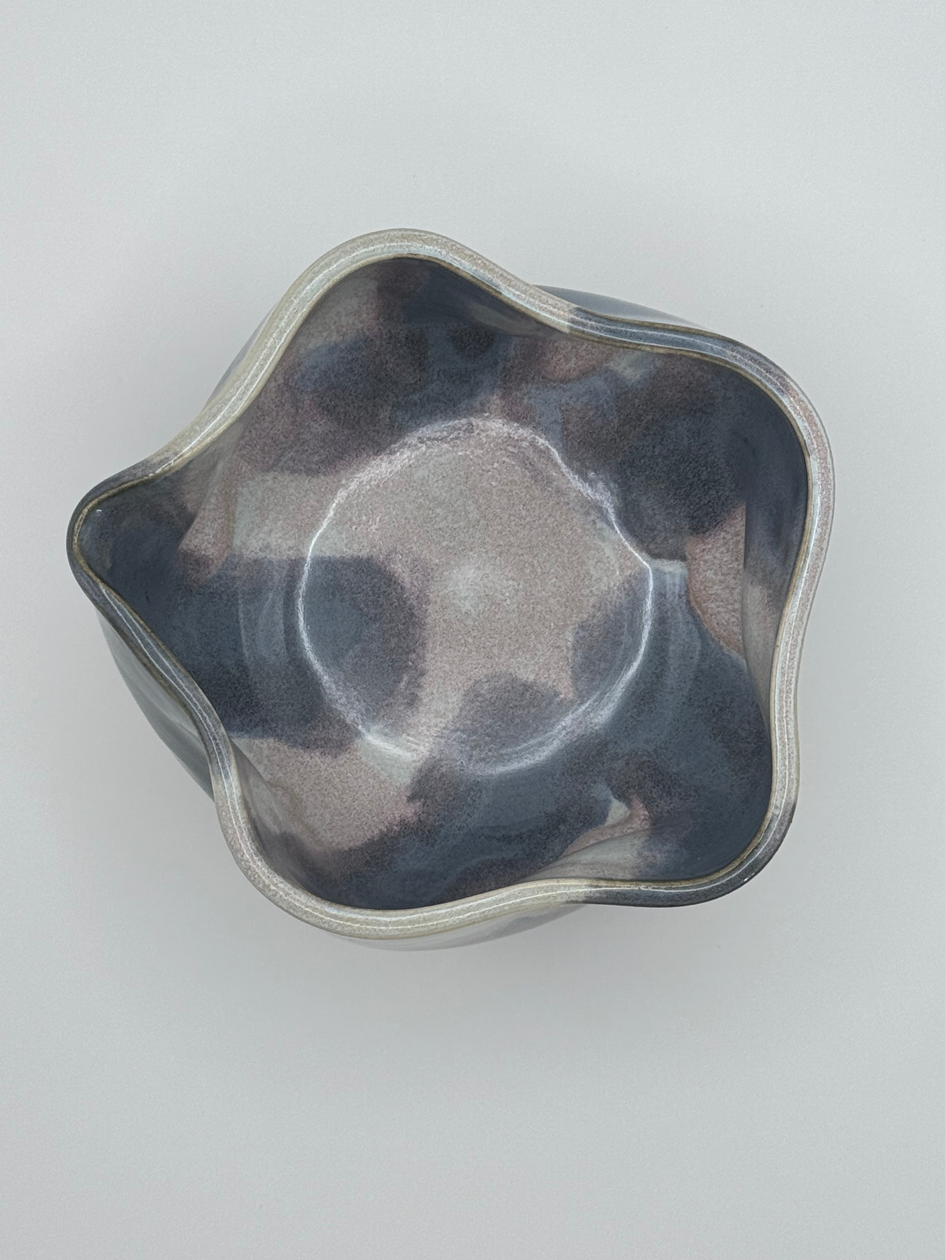 A top-down view of a handcrafted ceramic bowl with a wavy, scalloped rim and a marbled glaze in shades of slate blue, light blue, and soft dusty rose specks, set against a plain light gray background.