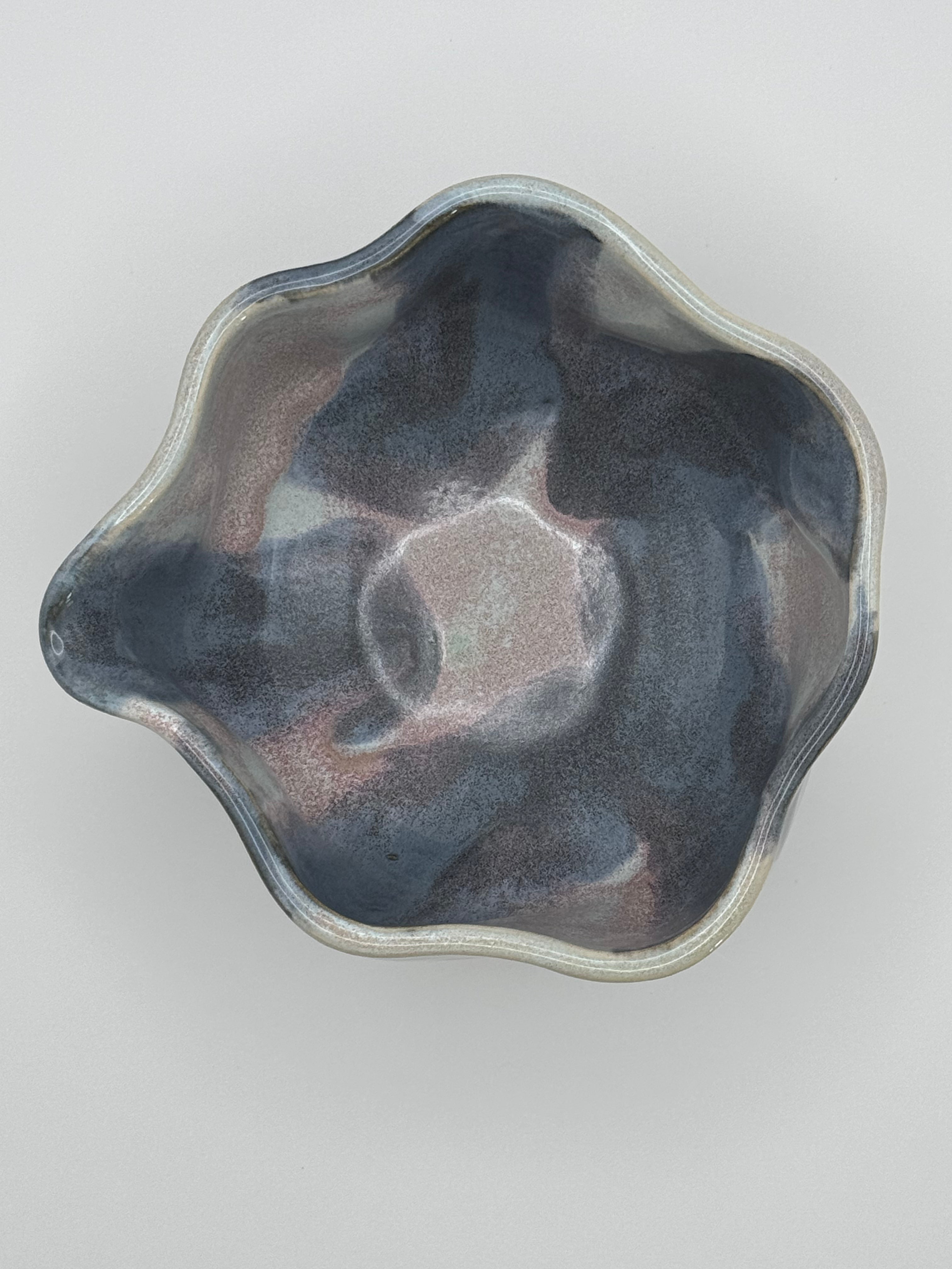 A handcrafted ceramic bowl with a scalloped, organic rim, featuring a glossy glaze with a marbled pattern of dark blue, light blue, and subtle pinkish tones specks, photographed from a top-down perspective on a neutral light gray background.