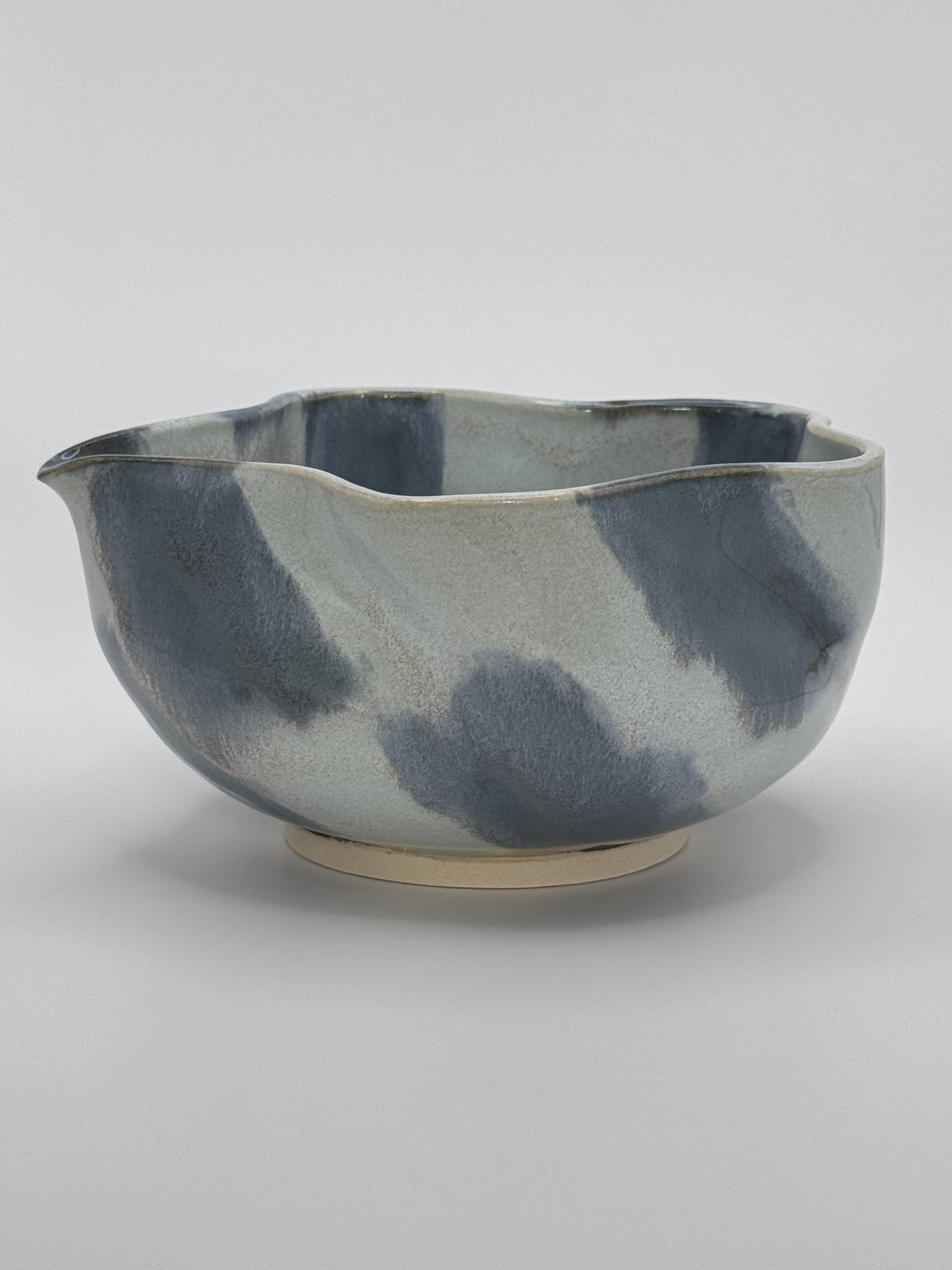 A handmade ceramic bowl with a wavy rim and a small footed base, featuring a glossy glaze with abstract slated blue and light blue brushstrokes over a white clay base, set against a plain light gray background.