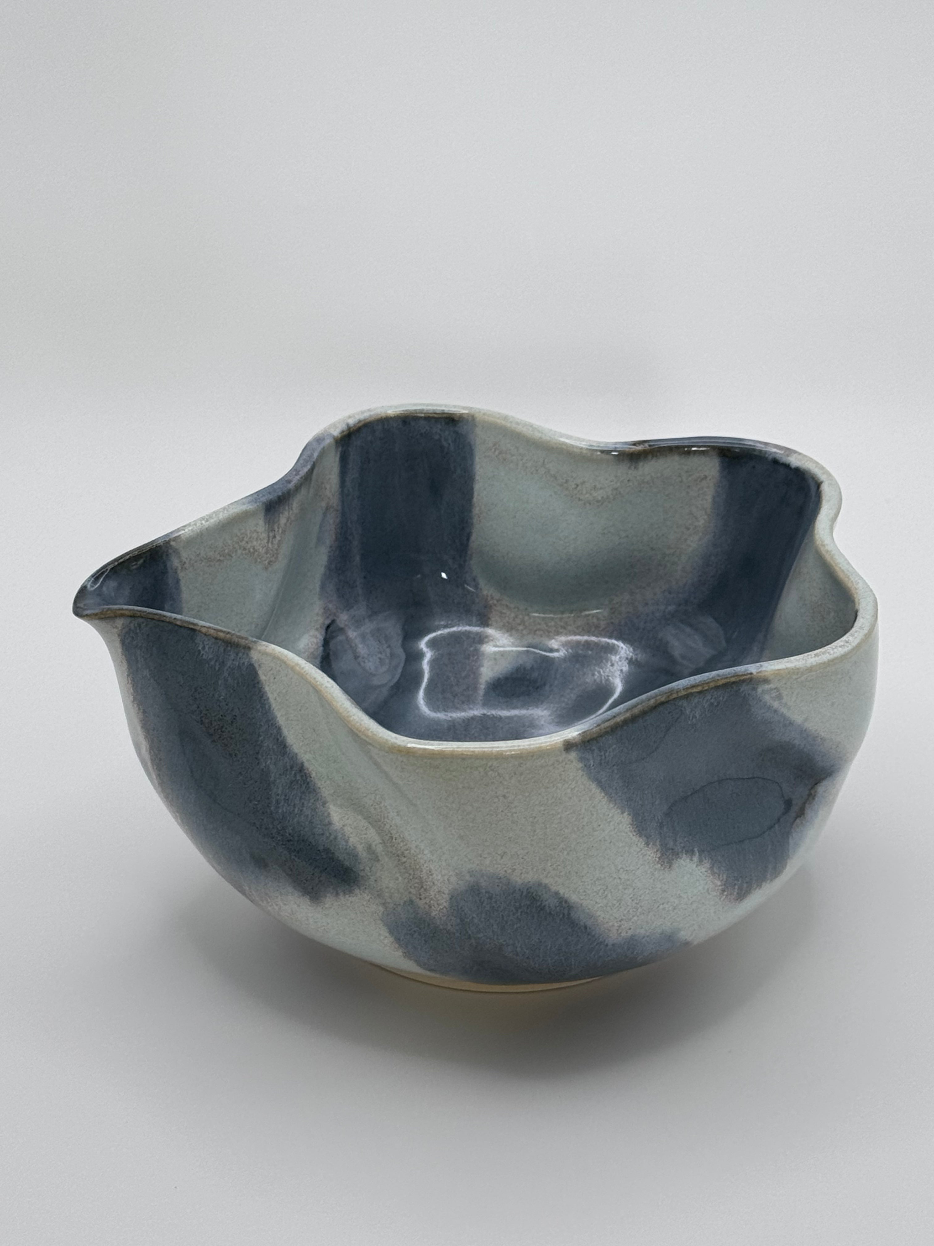 A handmade ceramic bowl with a wavy rim and a small footed base, featuring a glossy glaze with abstract slated blue and light blue brushstrokes over a white clay base, set against a plain light gray background.