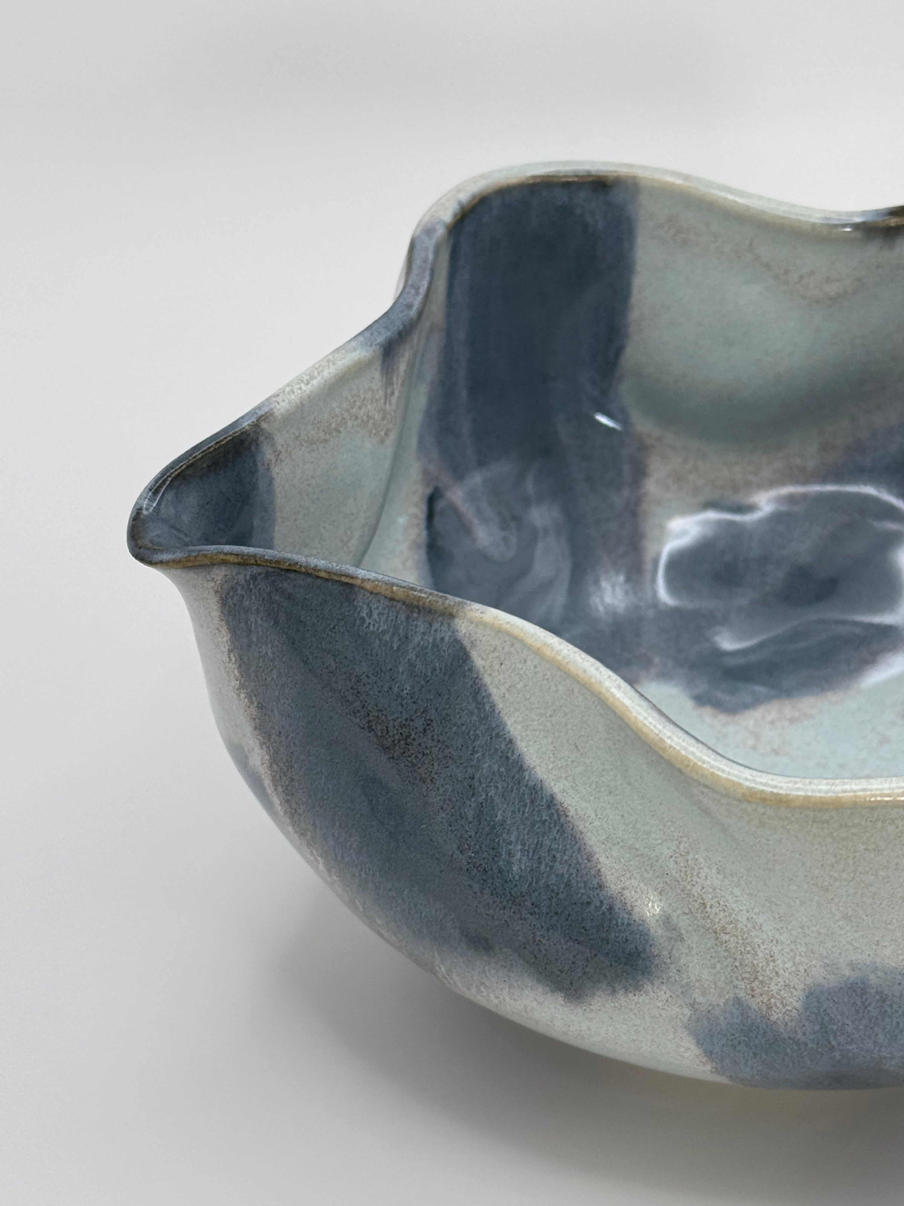 A close-up of A handmade ceramic bowl with a wavy rim and a small footed base, featuring a glossy glaze with abstract slated blue and light blue brushstrokes over a white clay base, set against a plain light gray background.