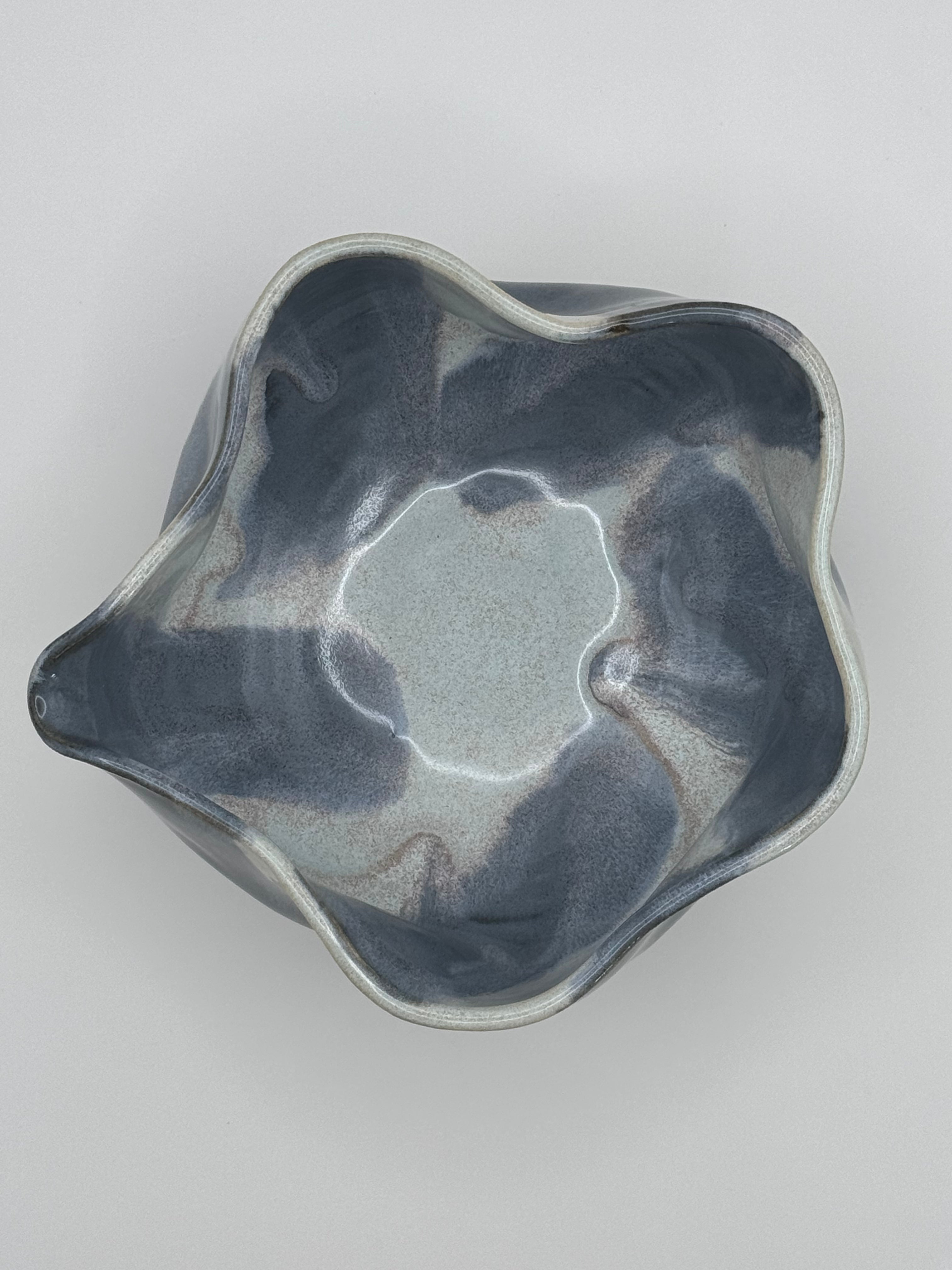 A handmade ceramic bowl with a wavy, scalloped rim, featuring a marbled glaze in shades of slate blue, light blue, and dusty rose specks, photographed from a top-down perspective on a neutral light gray background