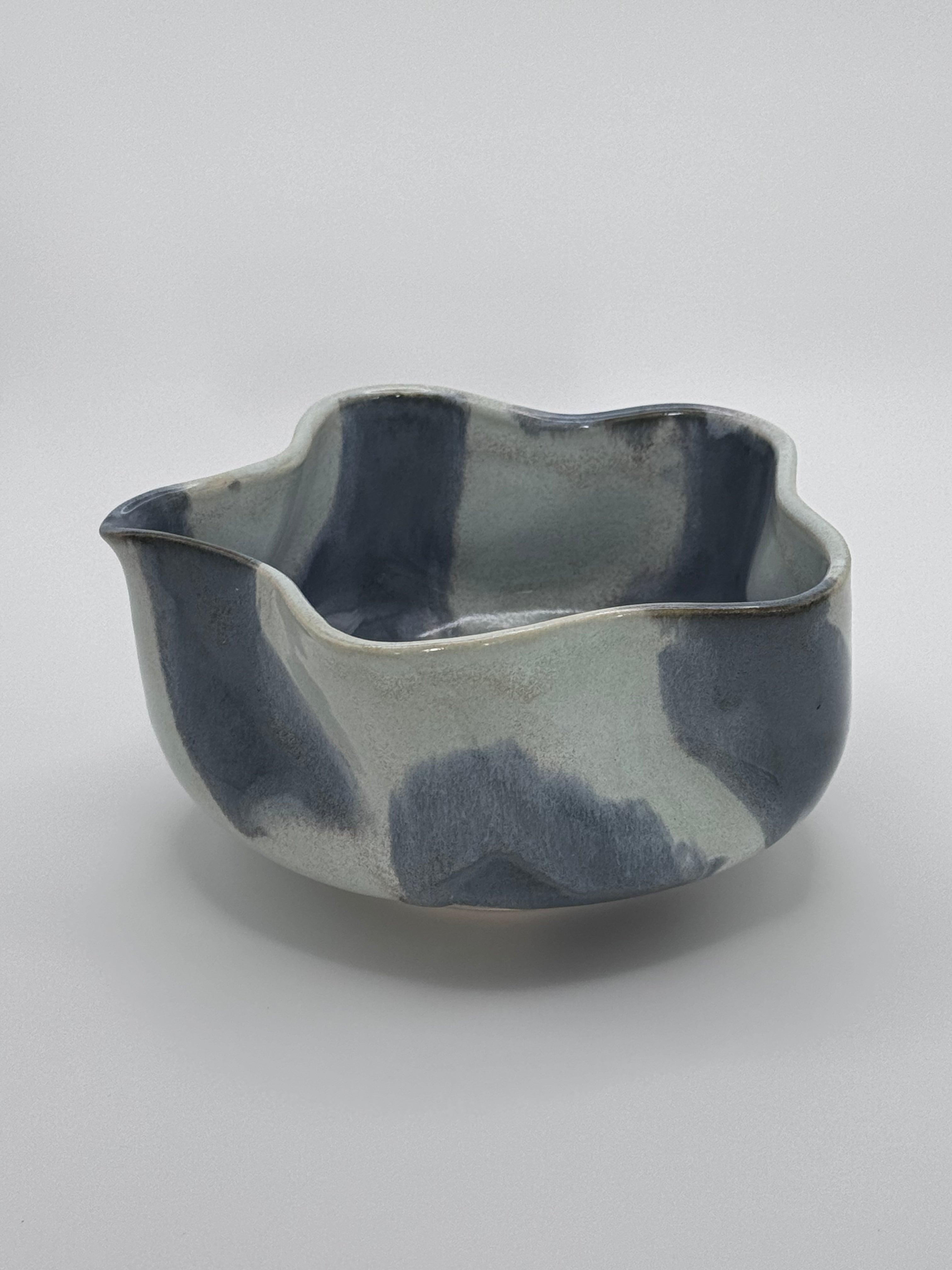 A handmade ceramic bowl with a wavy rim and a small footed base, featuring a glossy glaze with abstract slated blue and light blue brushstrokes over a white clay base, set against a plain light gray background.
