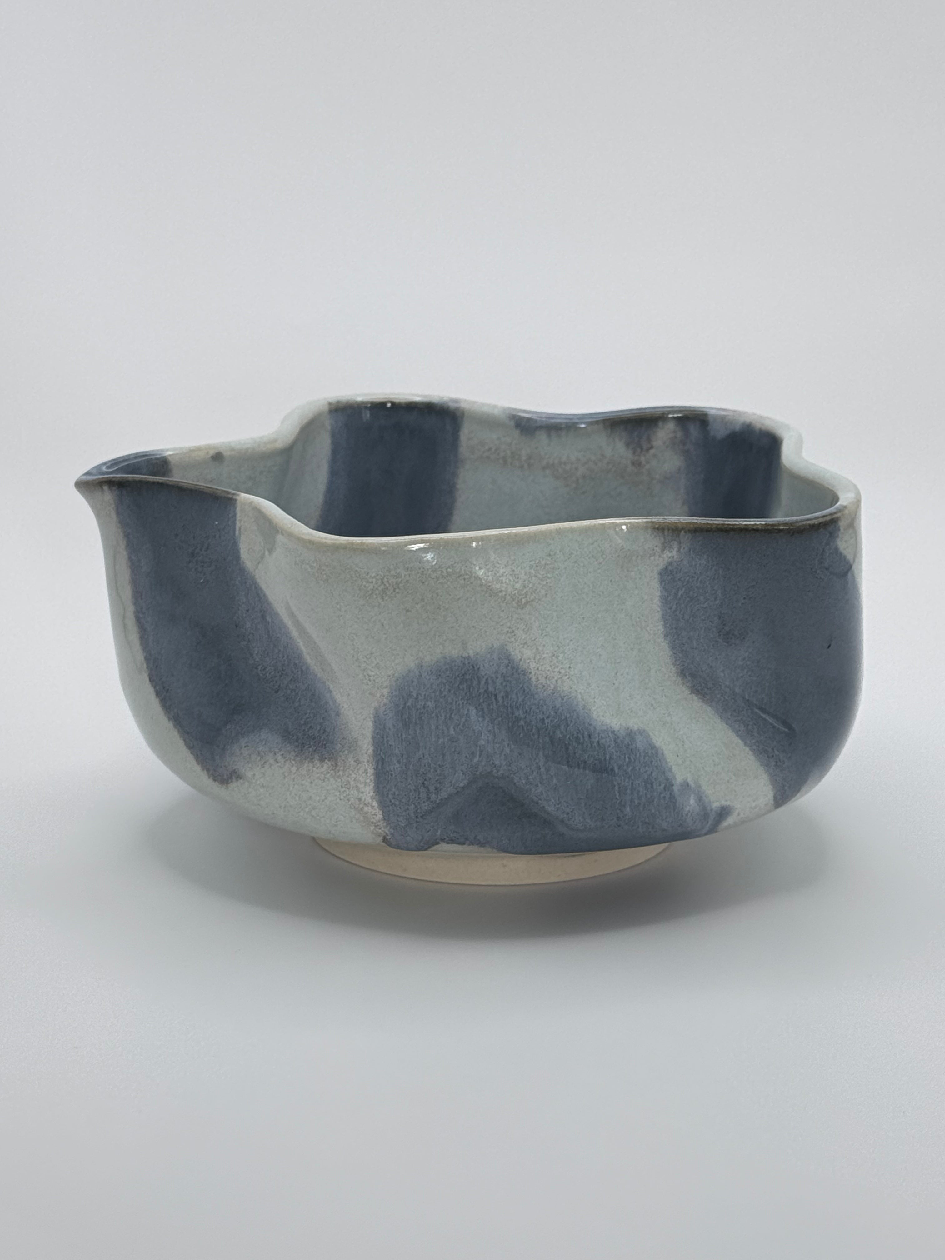 A handmade ceramic bowl with a wavy rim and a small footed base, featuring a glossy glaze with abstract slated blue and light blue brushstrokes over a white clay base, set against a plain light gray background.