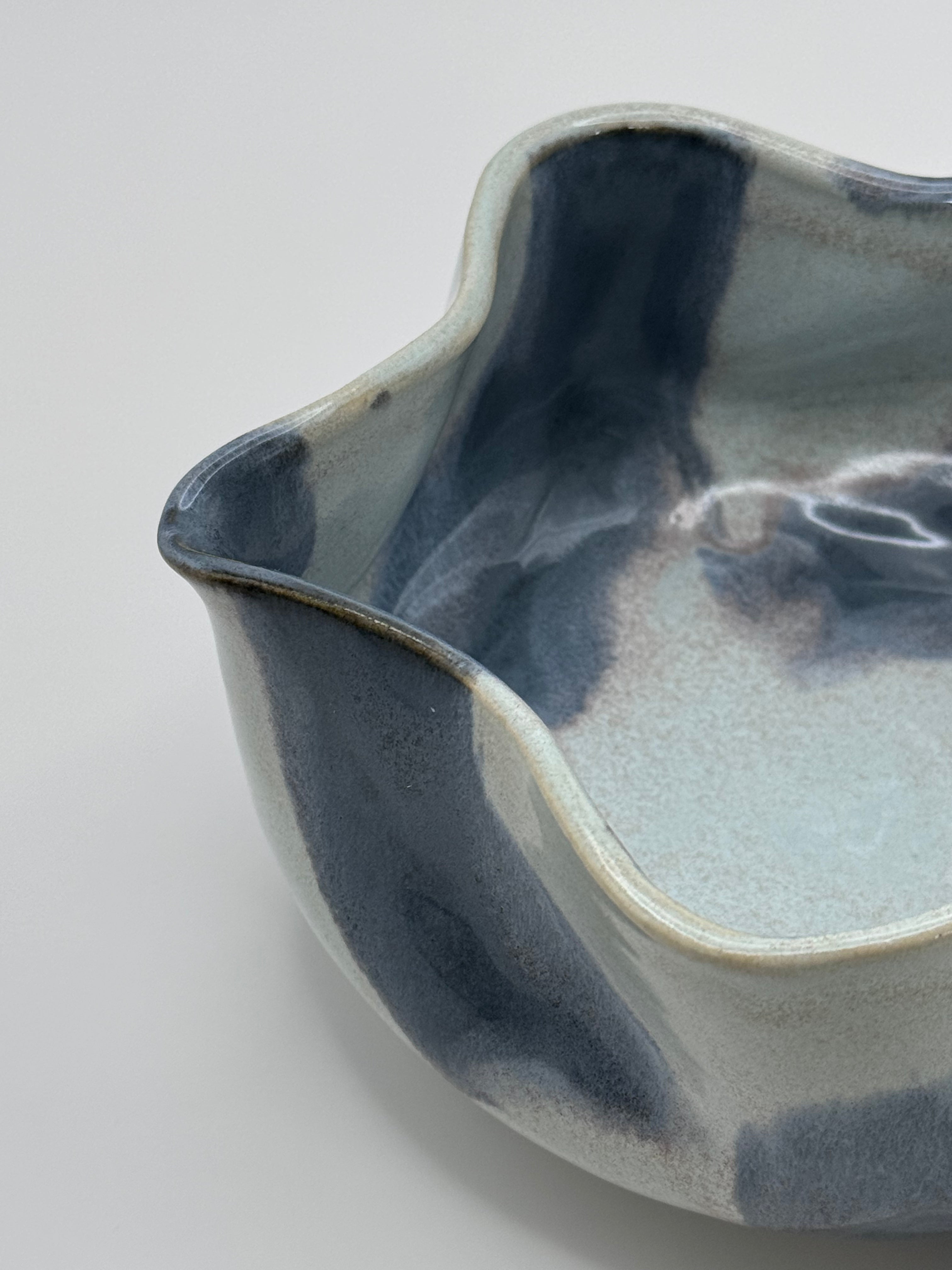 A close-up of A handmade ceramic bowl with a wavy rim and a small footed base, featuring a glossy glaze with abstract slated blue and light blue brushstrokes over a white clay base, set against a plain light gray background.
