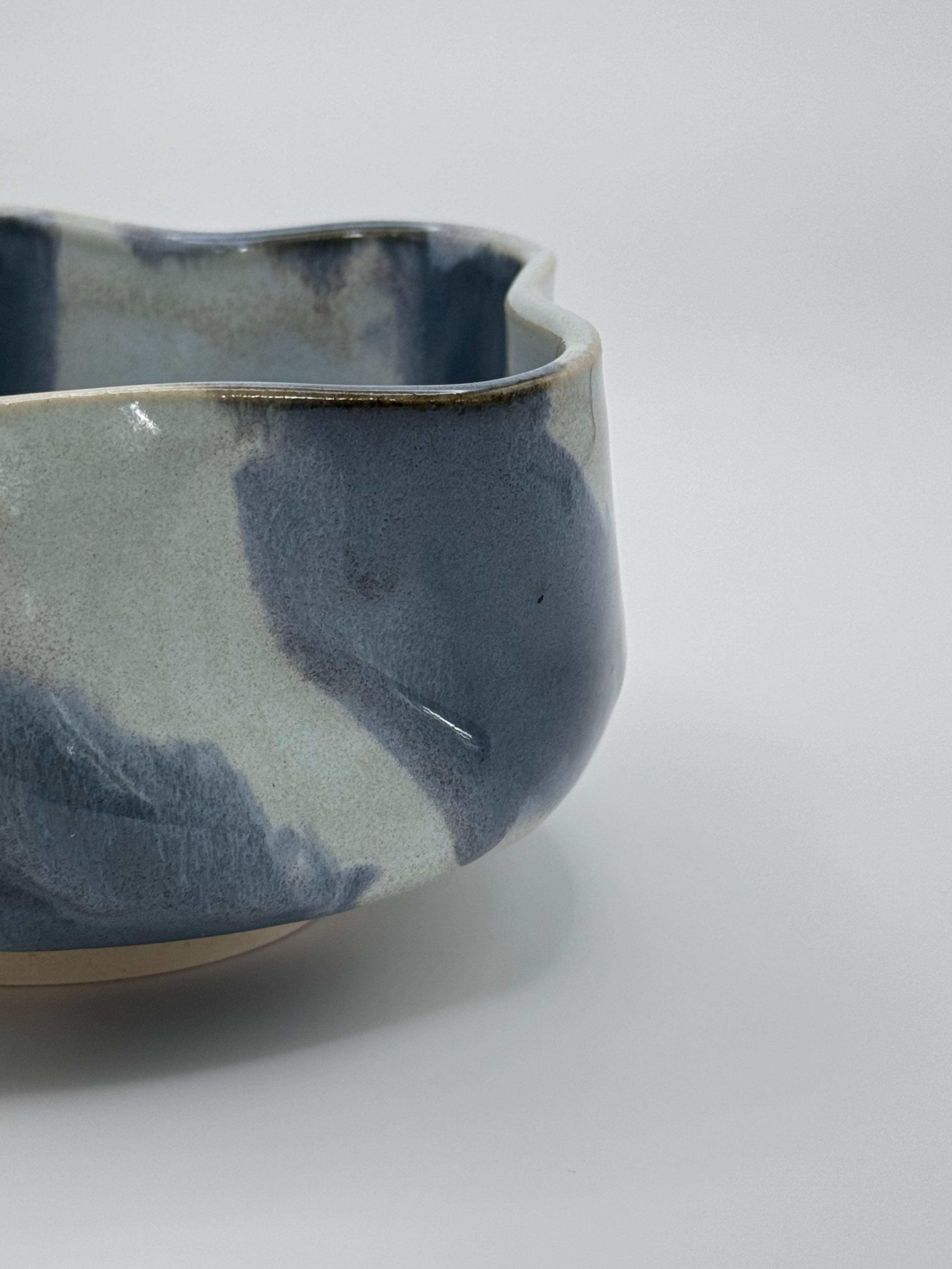 A close-up studio shot of A handmade ceramic bowl with a wavy rim and a small footed base, featuring a glossy glaze with abstract slated blue and light blue brushstrokes over a white clay base, set against a plain light gray background.