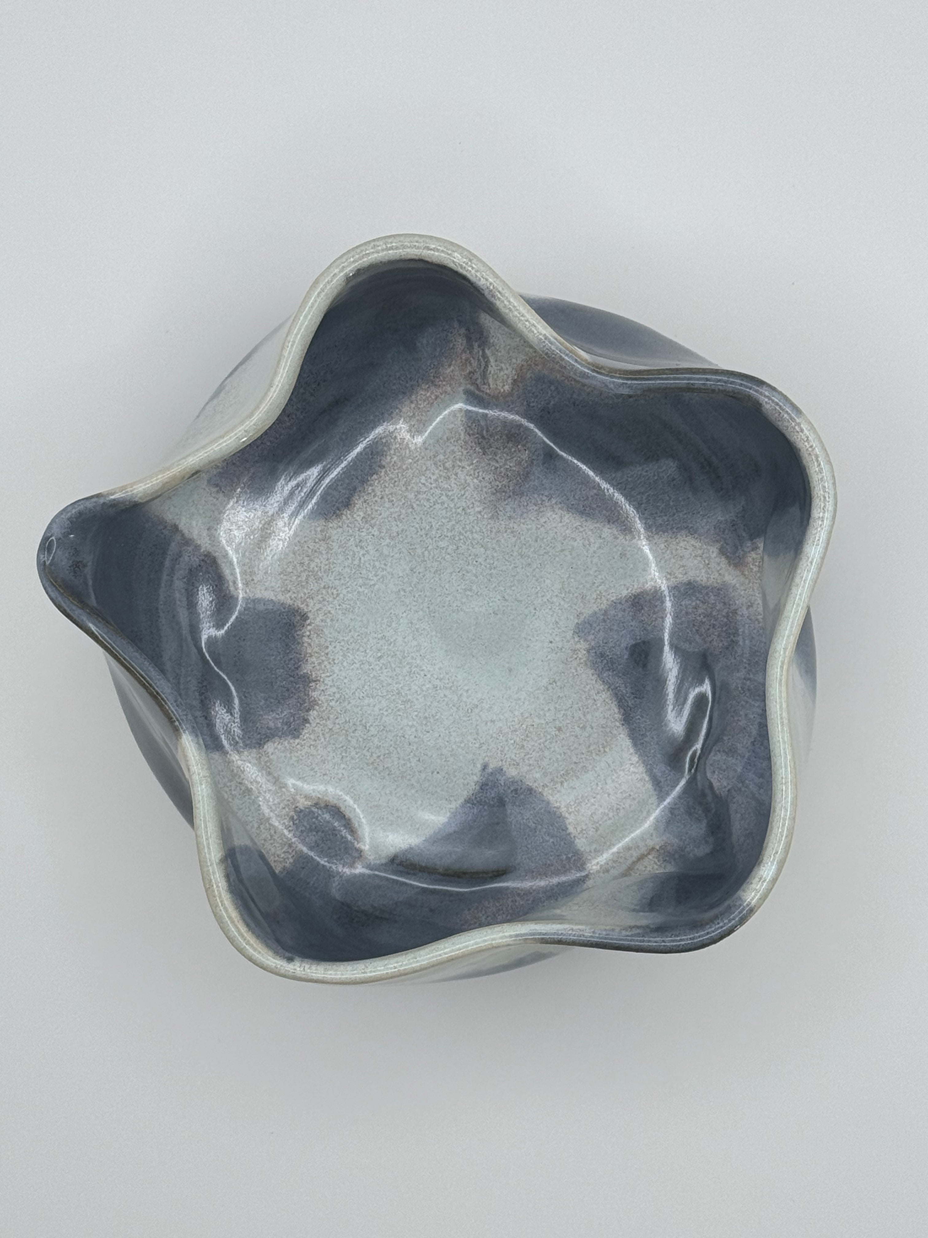 A ceramic bowl with a wavy, scalloped rim, featuring a glossy glaze with a gradient from dark slate blue-gray to a lighter blue interior with dusty rose specks, photographed from directly above on a neutral light gray background.